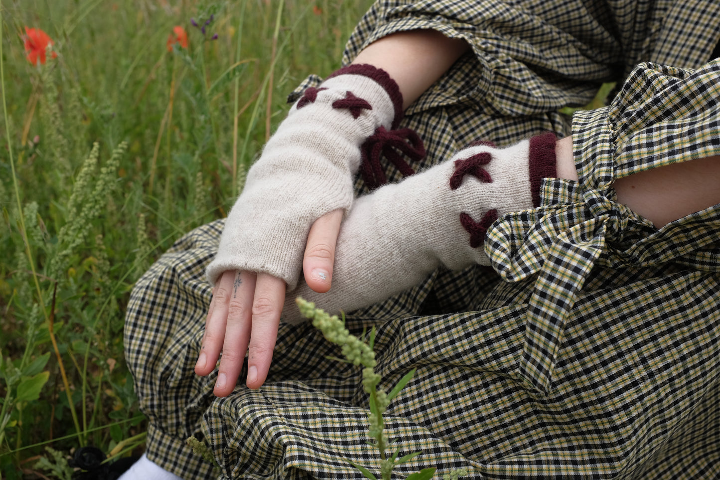 A woman sat in a filed, focused only on her hands wearing a paire of fingerless gloves in plum and cream. The base of the gloves is cream with the trim and tie detailing in a plum. The background of the image is the woman’s trendy, autumnal outfit.