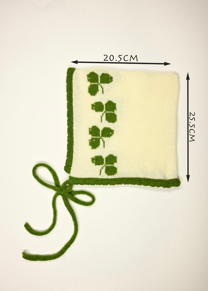 Green knitted bonnet with four-leaf clover patterns and measurements on a beige background