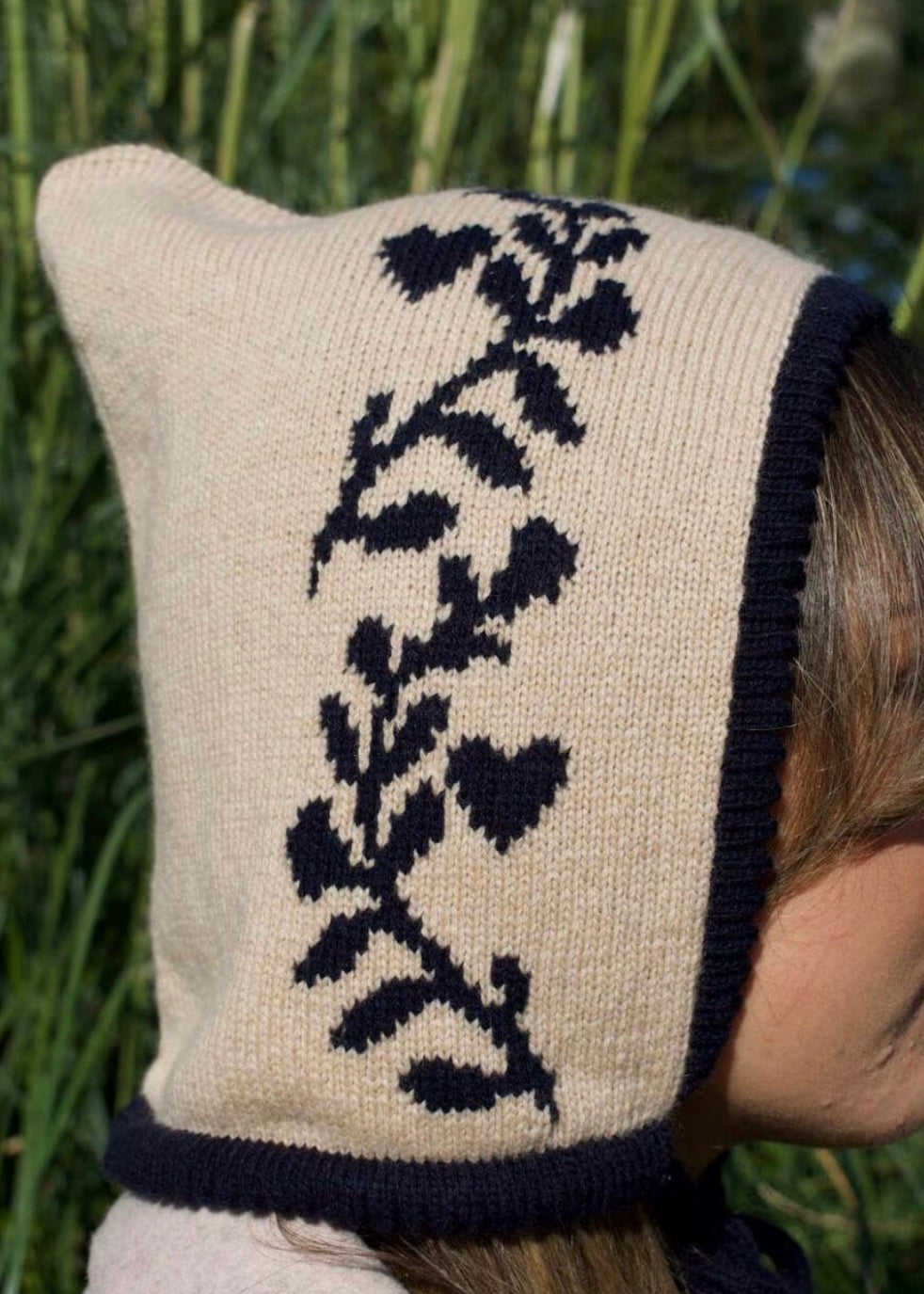 Knitted bonnet with navy floral pattern, trim and tie with a cream base, worn by a person in a natural setting