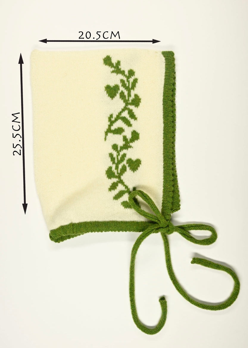Green knitted bonnet with floral pattern on a white background, shown with measurements of 20.5cm wide by 25.5cm long