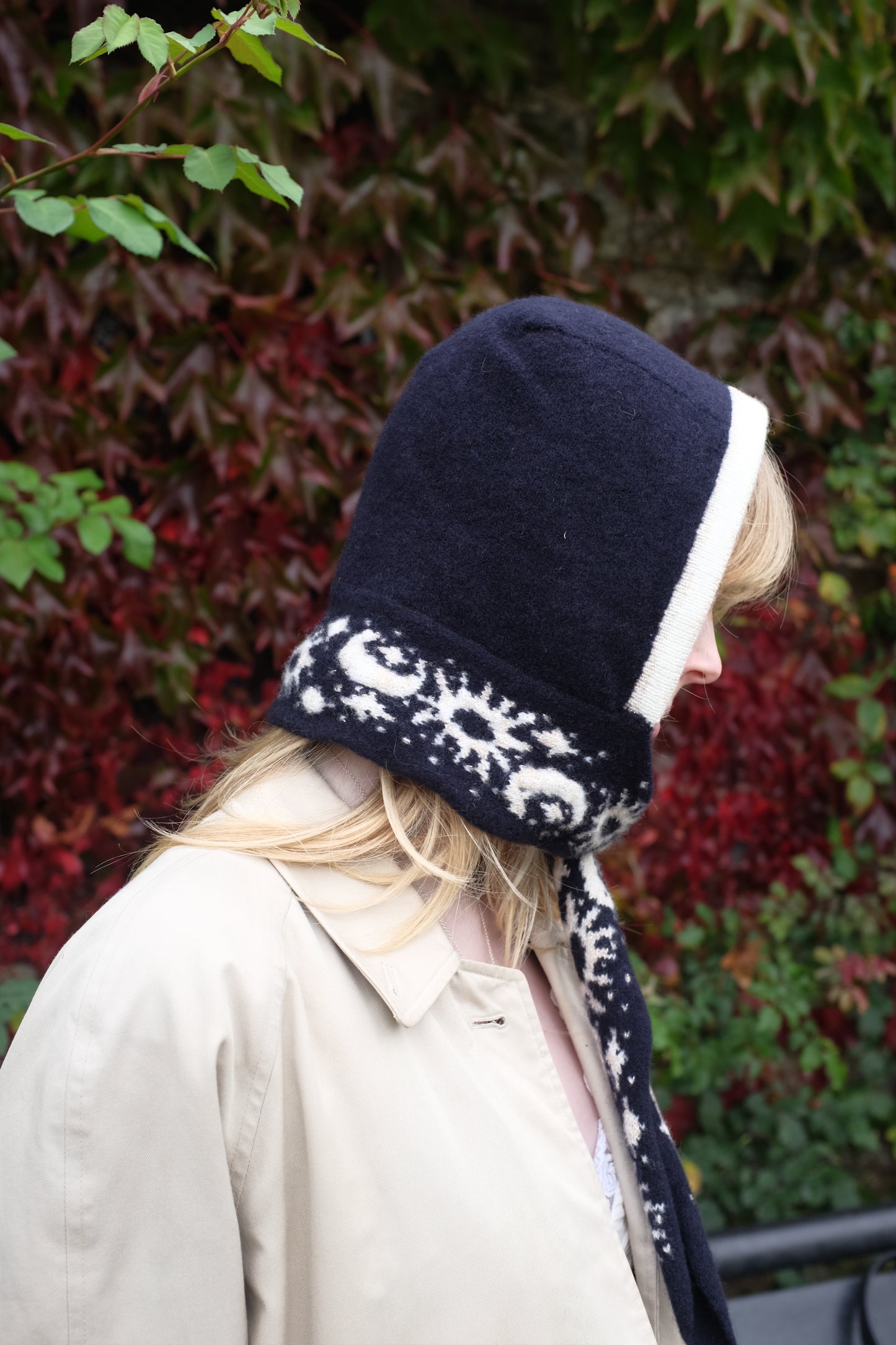 A women looking to the side wearing a lambswool hand crafted bonnet, with a scarf detailing around the bottom. The main colour of the bonnet is navy. With a white trim, and around the scarf detailing there is a celestial print of the moon and stars in white with gold shimmering details. Behind the women is a blurry mix of green and red leaves 