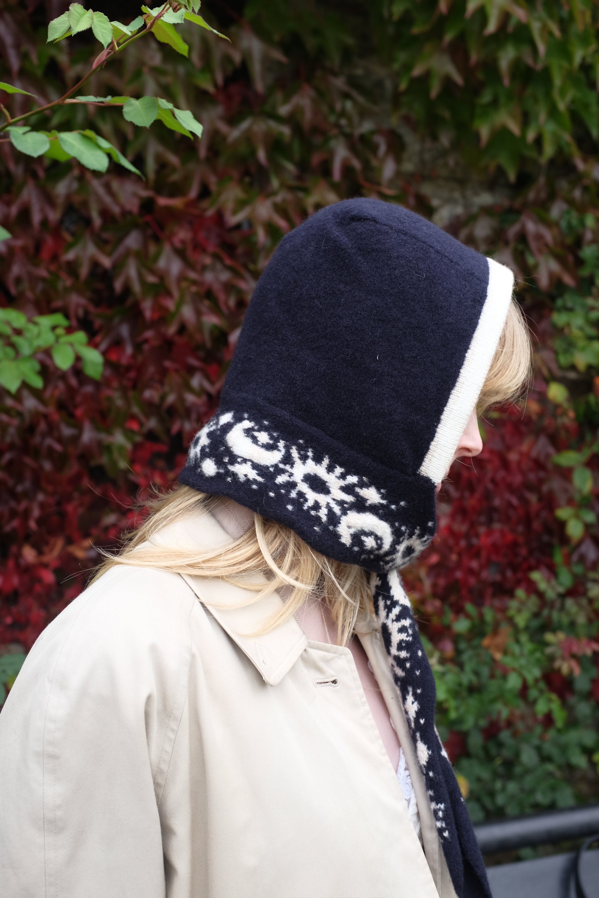 A women looking to the side wearing a lambswool hand crafted bonnet, with a scarf detailing around the bottom. The main colour of the bonnet is navy. With a white trim, and around the scarf detailing there is a celestial print of the moon and stars in white with gold shimmering details. Behind the women is a blurry mix of green and red leaves 