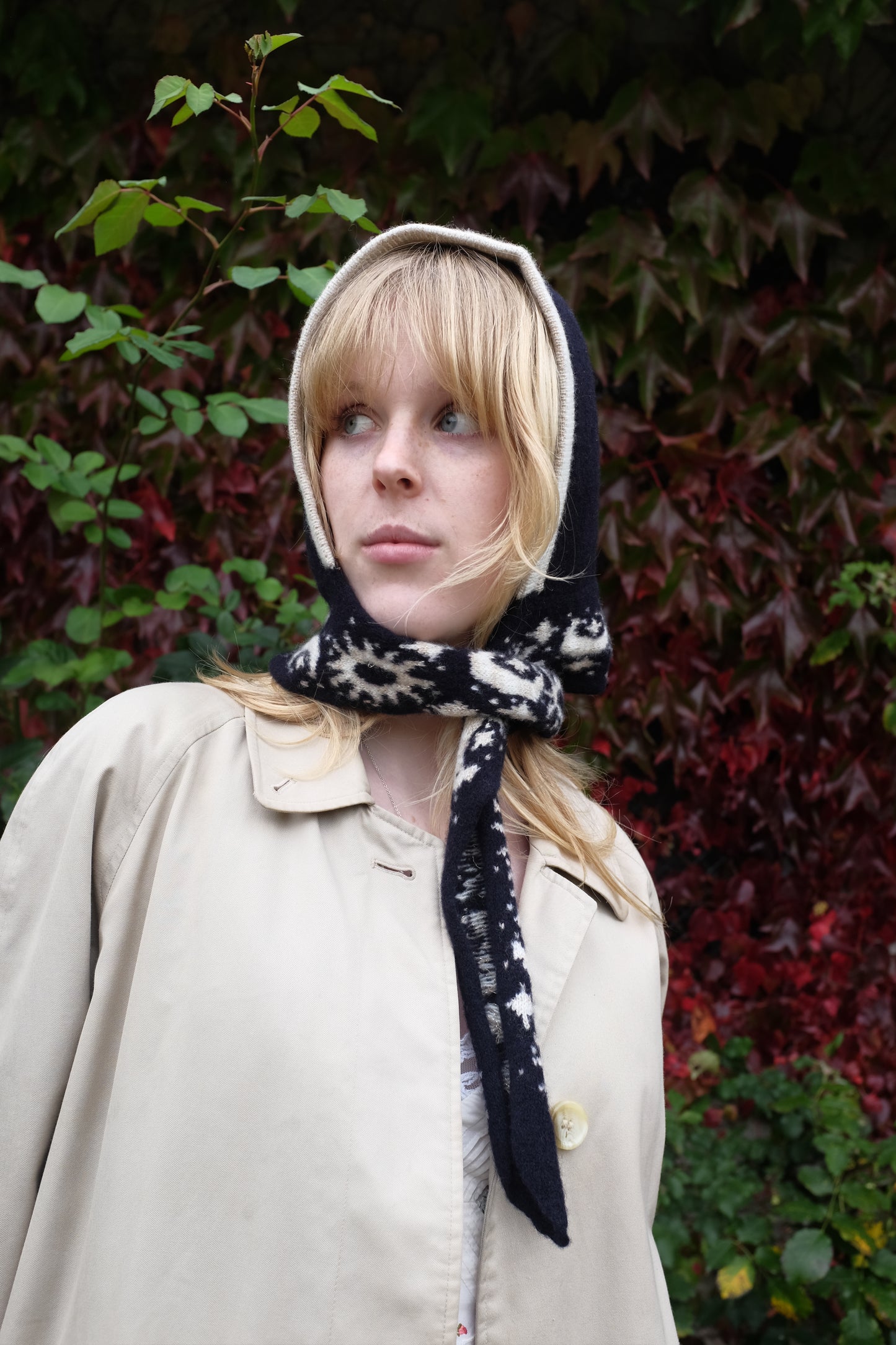 A women looking to the side wearing a lambswool hand crafted bonnet, with a scarf detailing around the bottom. The main colour of the bonnet is navy. With a white trim, and around the scarf detailing there is a celestial print of the moon and stars in white with gold shimmering details. Behind the women is a blurry mix of green and red leaves 