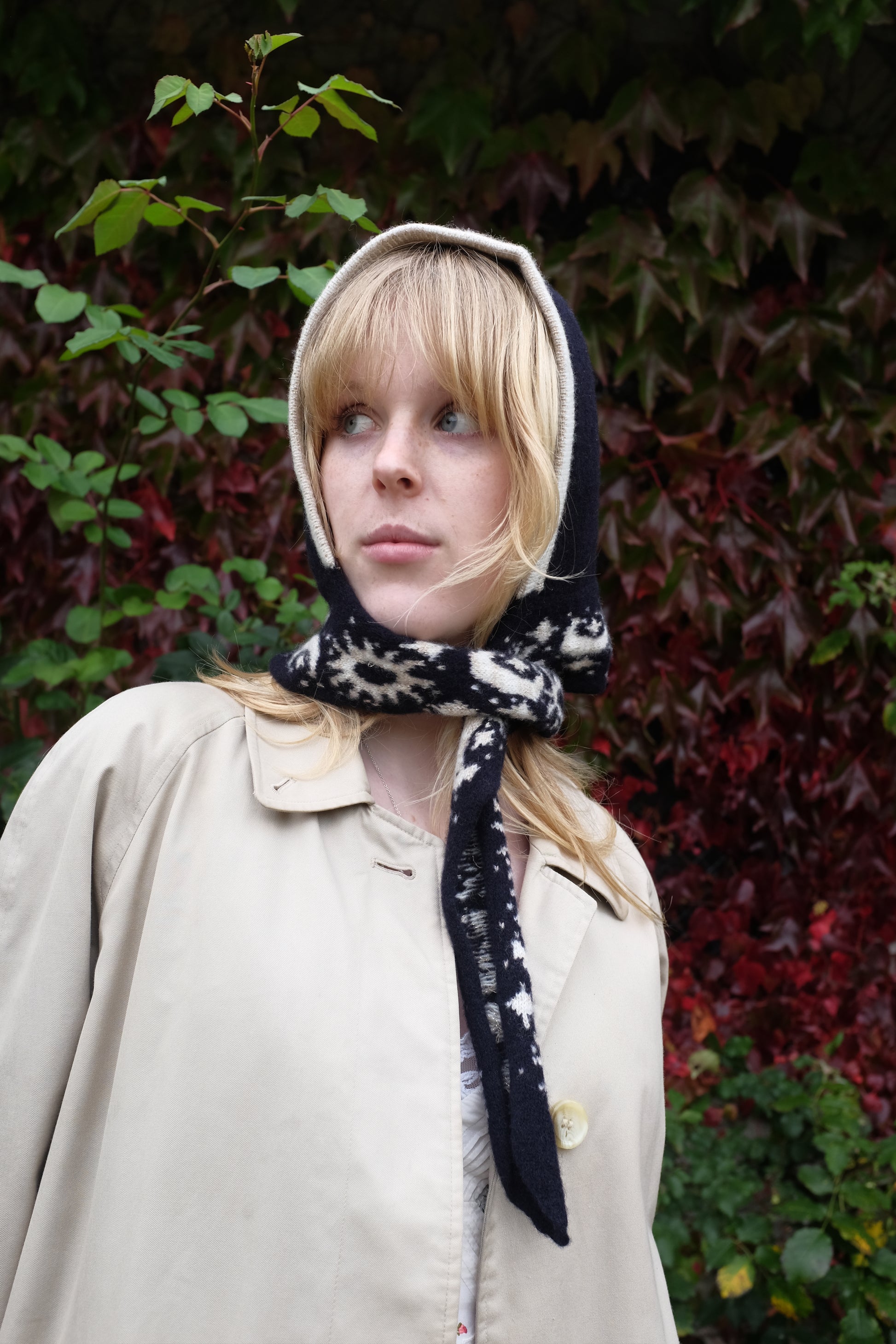 A women looking to the side wearing a lambswool hand crafted bonnet, with a scarf detailing around the bottom. The main colour of the bonnet is navy. With a white trim, and around the scarf detailing there is a celestial print of the moon and stars in white with gold shimmering details. Behind the women is a blurry mix of green and red leaves 