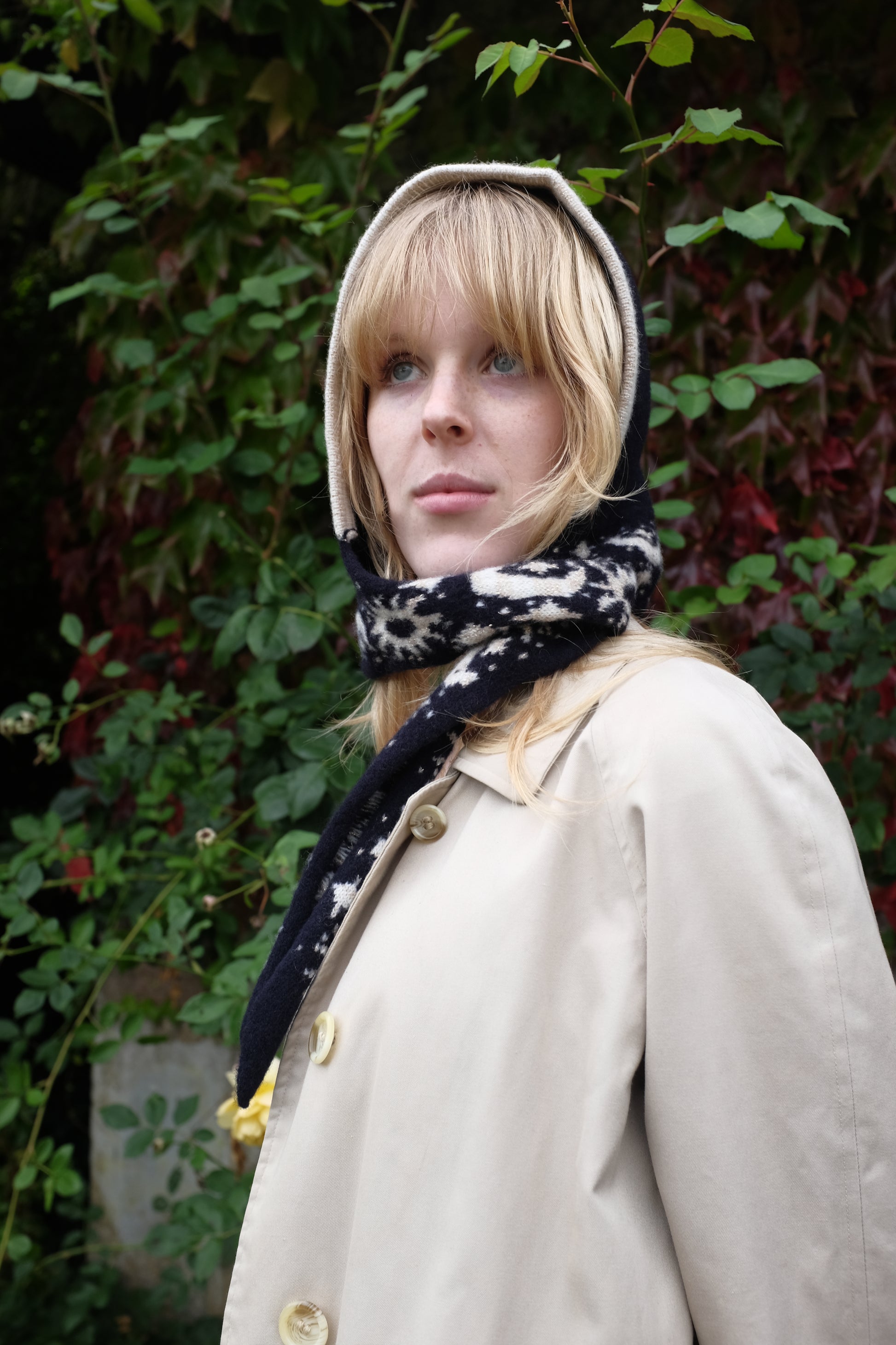 A women looking to the side wearing a lambswool hand crafted bonnet, with a scarf detailing around the bottom. The main colour of the bonnet is navy. With a white trim, and around the scarf detailing there is a celestial print of the moon and stars in white with gold shimmering details. Behind the women is a blurry mix of green and red leaves 