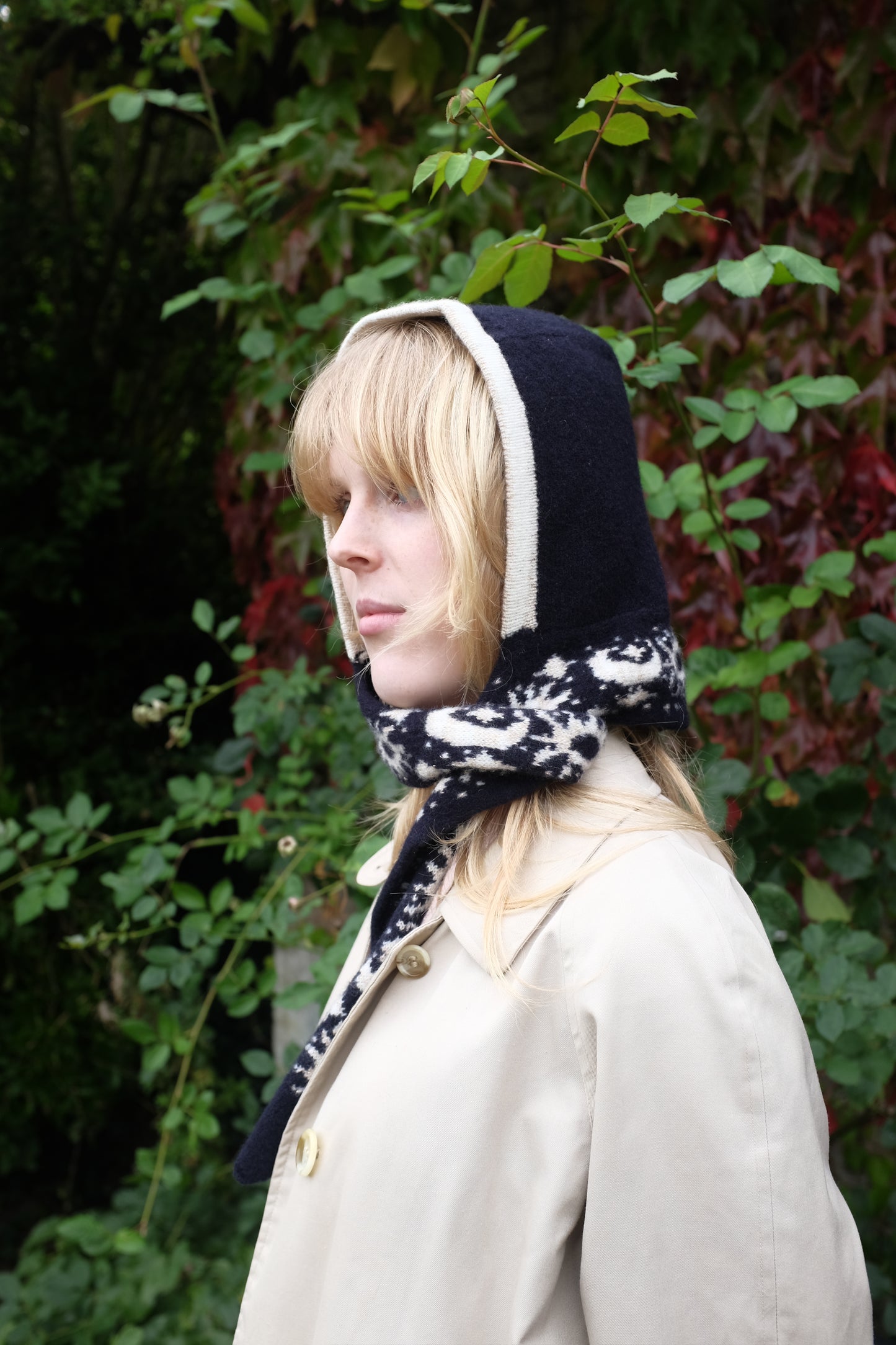 A women looking to the side wearing a lambswool hand crafted bonnet, with a scarf detailing around the bottom. The main colour of the bonnet is navy. With a white trim, and around the scarf detailing there is a celestial print of the moon and stars in white with gold shimmering details. Behind the women is a blurry mix of green and red leaves 