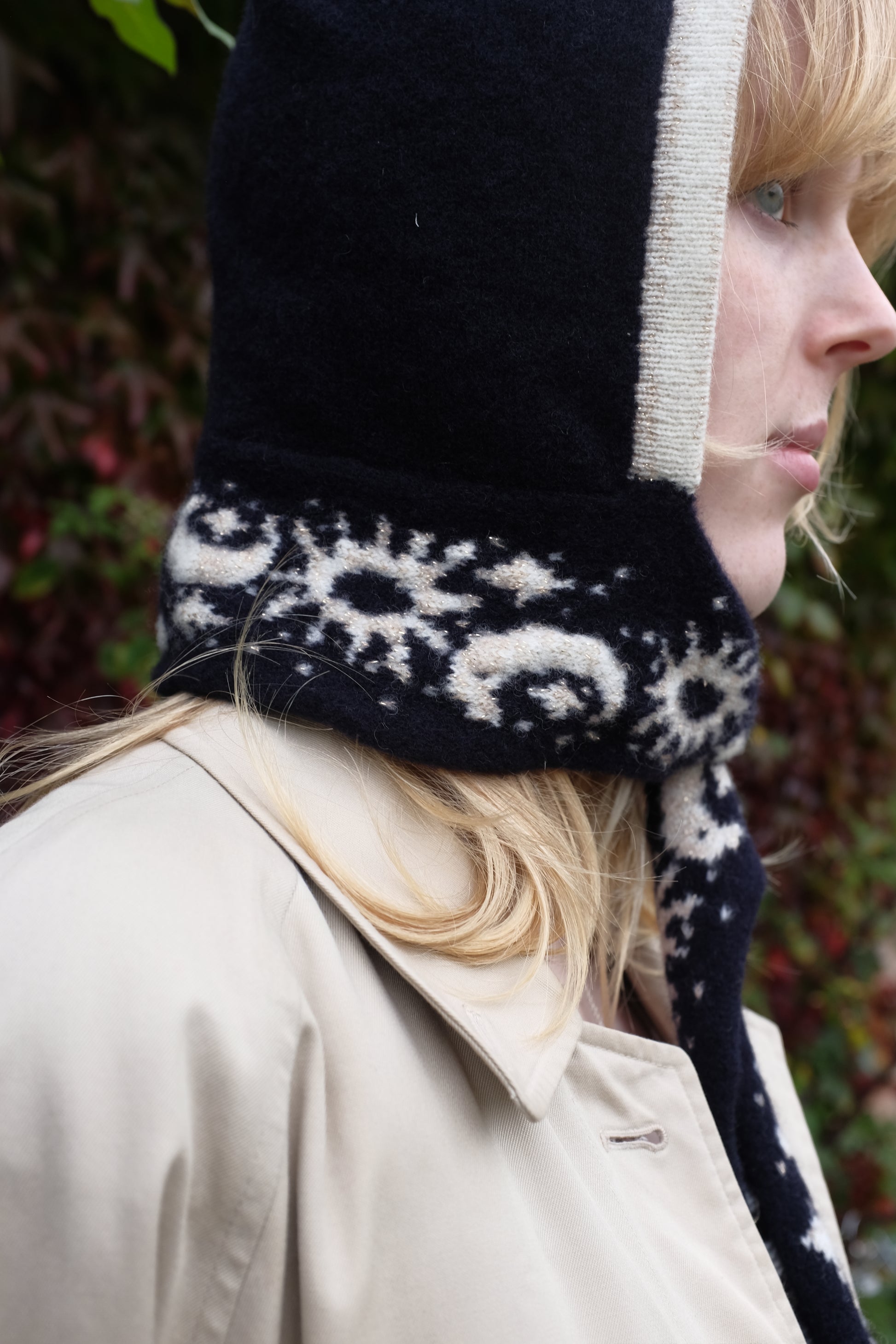 A close up of a women looking to the side wearing a lambswool hand crafted bonnet, with a scarf detailing around the bottom. The main colour of the bonnet is navy. With a white trim, and around the scarf detailing there is a celestial print of the moon and stars in white with gold shimmering details. Behind the women is a blurry mix of green and red leaves 
