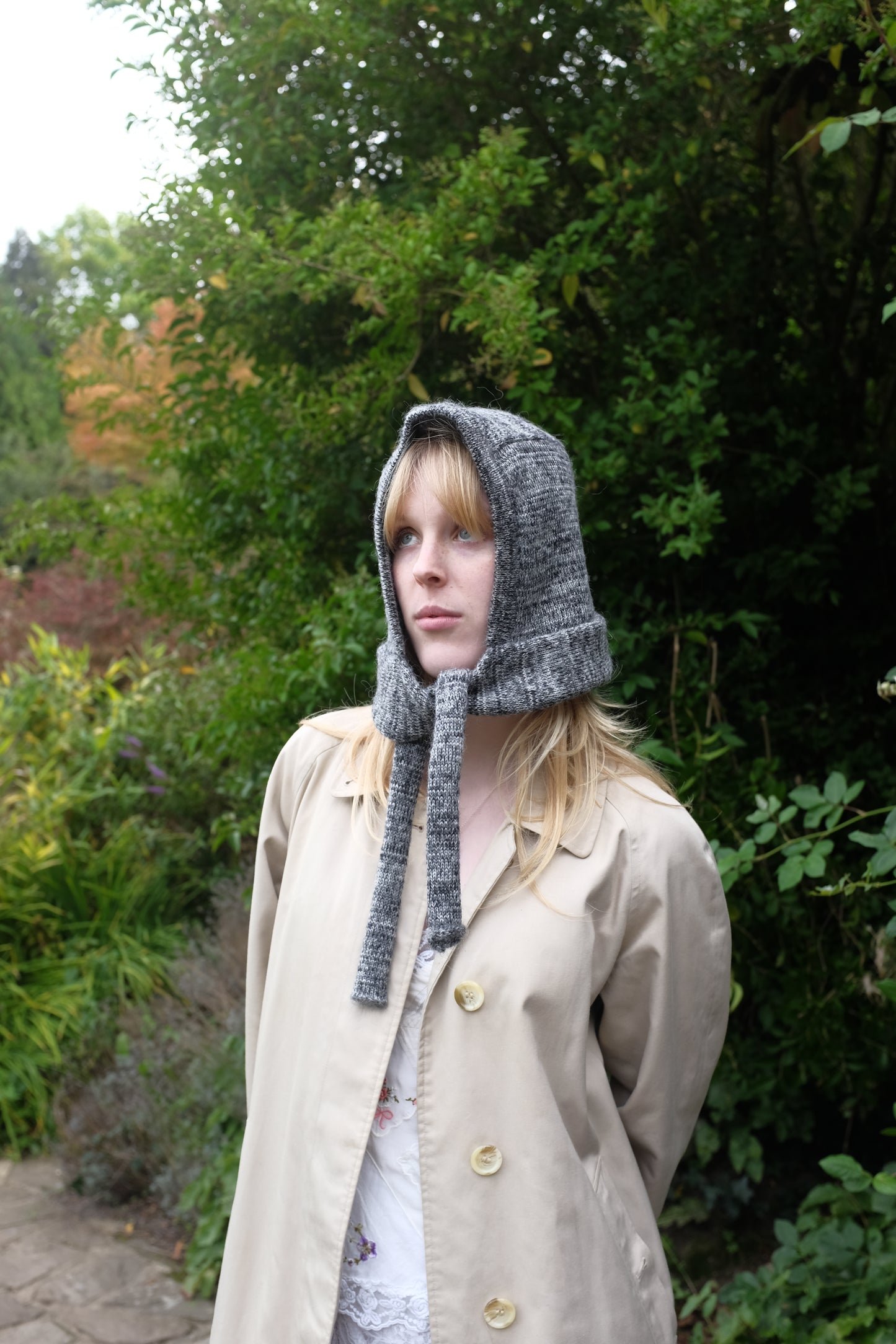 Person wearing a black mohair bonnet-style hood that fully covers the head and neck. With two long, soft extensions hanging down from the front. The hood is thick and textured, offering warmth and a cosy appearance, ideal for cold weather. The person is dressed in a light coloured coat and stands outdoors against a backdrop of green and red foliage 