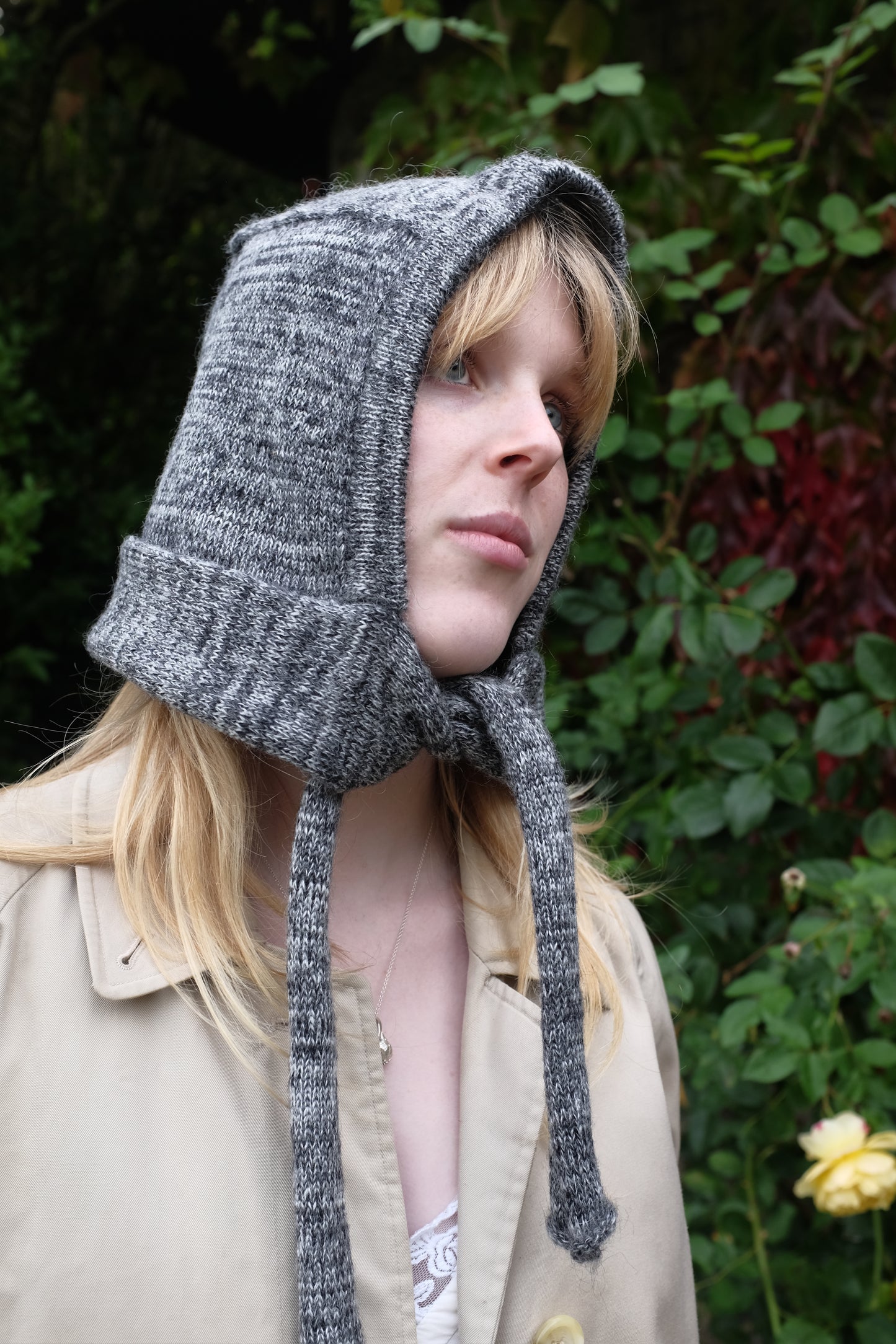 Person wearing a black mohair bonnet-style hood that fully covers the head and neck. With two long, soft extensions hanging down from the front. The hood is thick and textured, offering warmth and a cosy appearance, ideal for cold weather. The person is dressed in a light coloured coat and stands outdoors against a backdrop of green and red foliage 