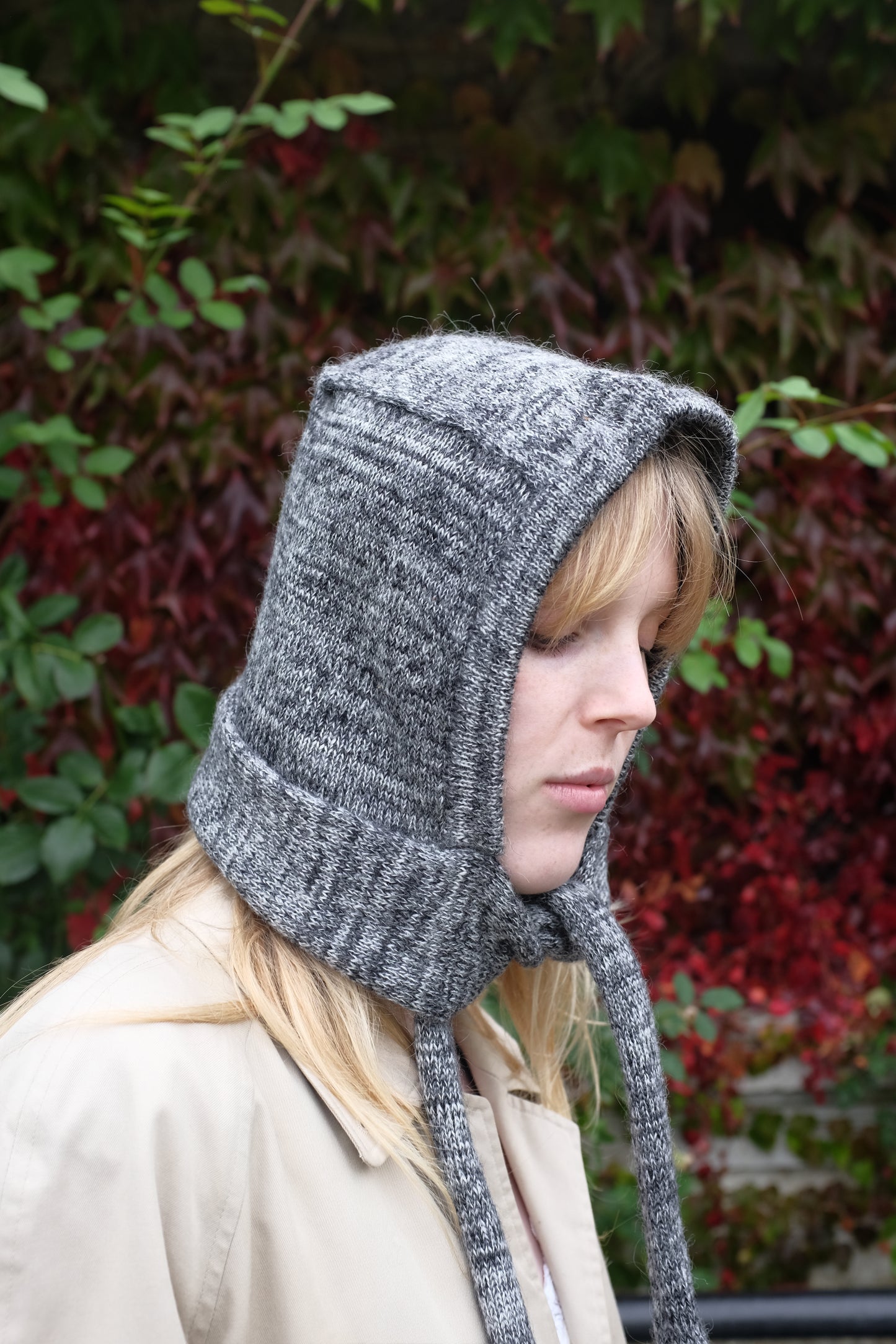 Person wearing a black mohair bonnet-style hood that fully covers the head and neck. With two long, soft extensions hanging down from the front. The hood is thick and textured, offering warmth and a cosy appearance, ideal for cold weather. The person is dressed in a light coloured coat and stands outdoors against a backdrop of green and red foliage 