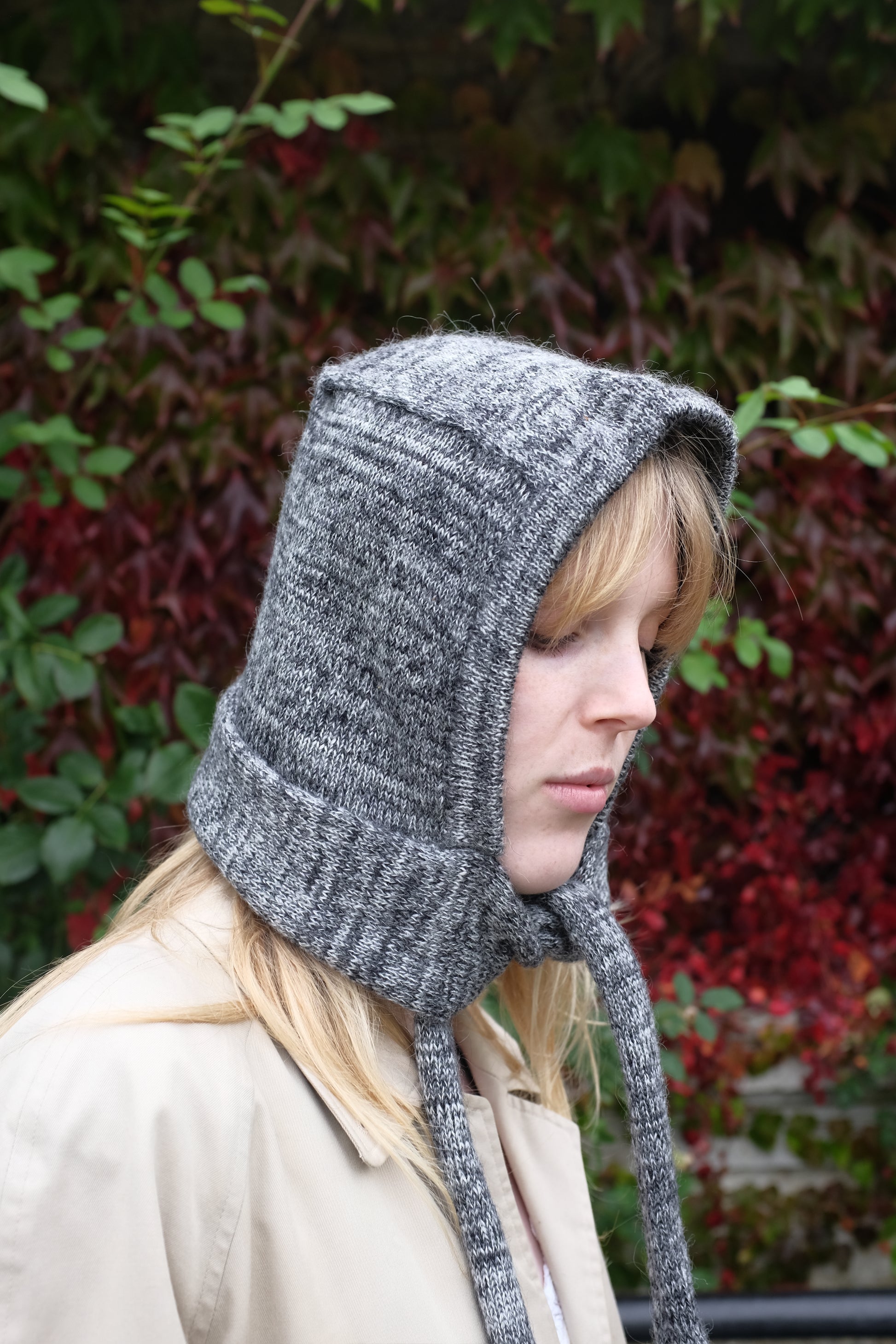 Person wearing a black mohair bonnet-style hood that fully covers the head and neck. With two long, soft extensions hanging down from the front. The hood is thick and textured, offering warmth and a cosy appearance, ideal for cold weather. The person is dressed in a light coloured coat and stands outdoors against a backdrop of green and red foliage 