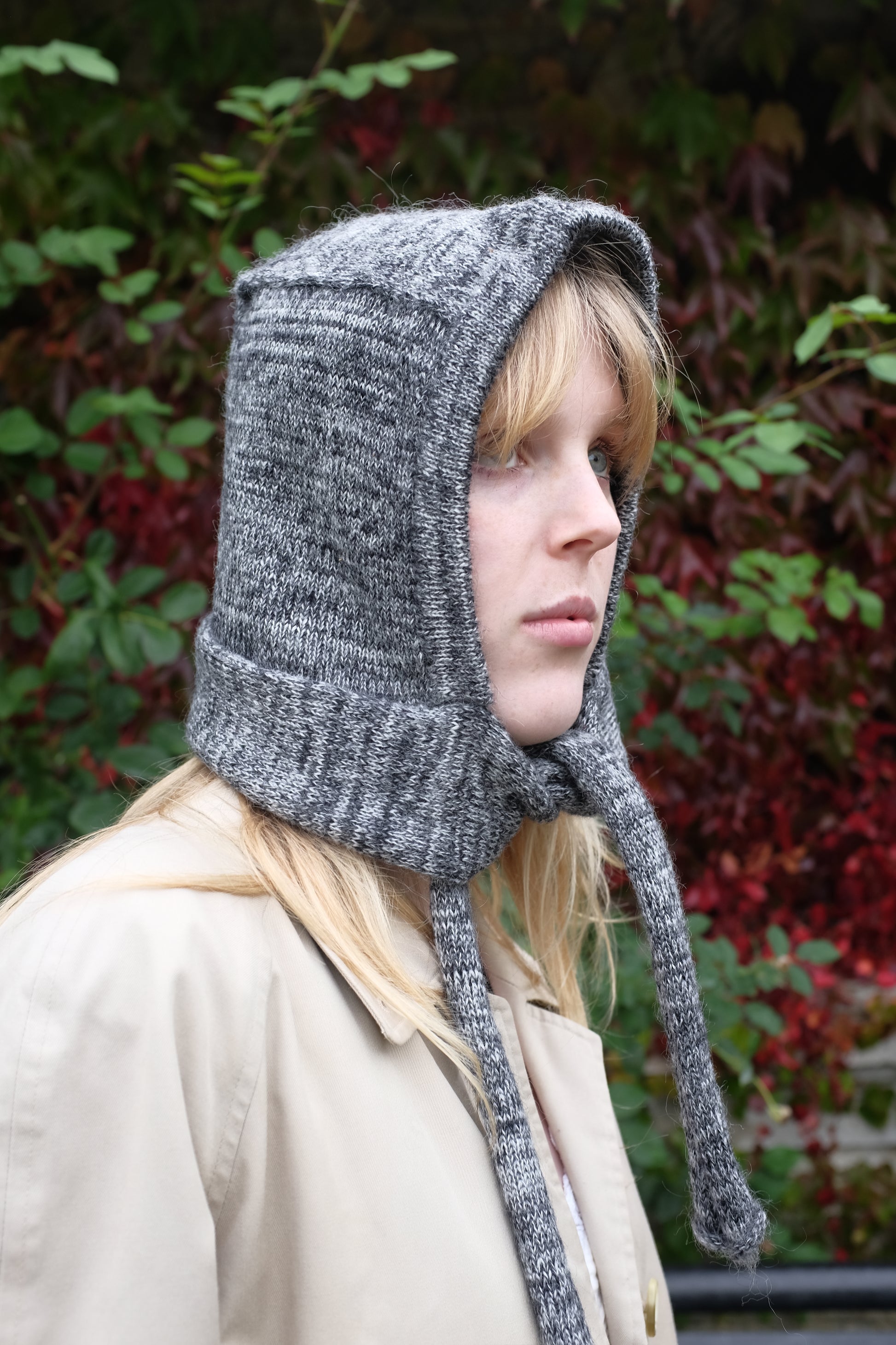 Person wearing a black mohair bonnet-style hood that fully covers the head and neck. With two long, soft extensions hanging down from the front. The hood is thick and textured, offering warmth and a cosy appearance, ideal for cold weather. The person is dressed in a light coloured coat and stands outdoors against a backdrop of green and red foliage 