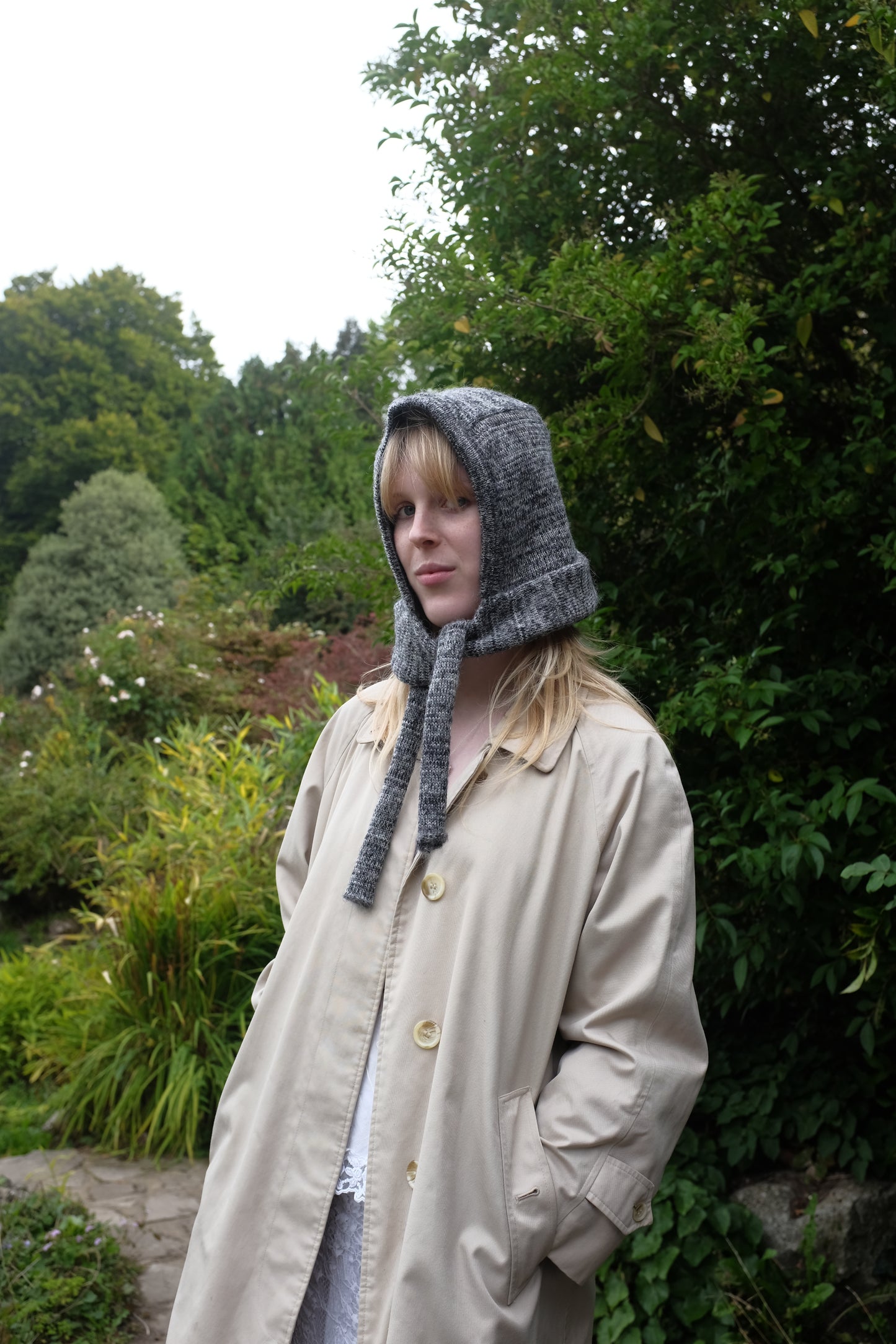 Person wearing a black mohair bonnet-style hood that fully covers the head and neck. With two long, soft extensions hanging down from the front. The hood is thick and textured, offering warmth and a cosy appearance, ideal for cold weather. The person is dressed in a light coloured coat and stands outdoors against a backdrop of green and red foliage 