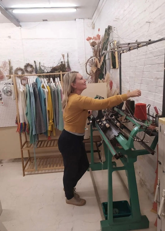 Person interacting with a large knit machine machine in a room with clothing racks and various items on the wall.