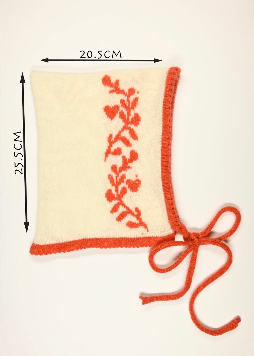 Cream cashmere knitted bonnet with an orange laurel leaf design and a tied strap on a white background, displaying the measurements of 20.5cm wide by 25.5cm long