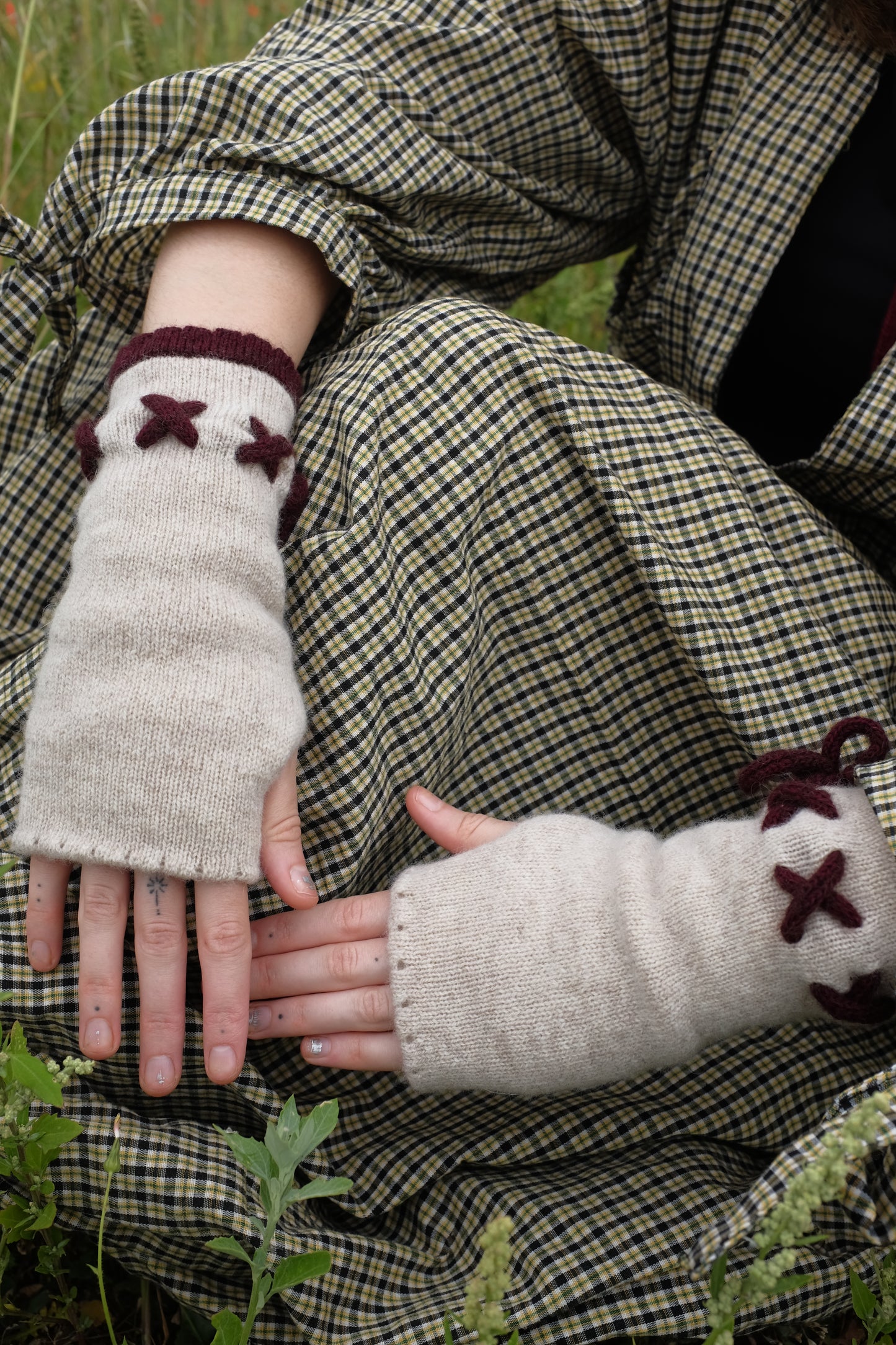 A woman sat in a filed, focuses only on her hands wearing a pair of fingerless gloves in plum and cream. The base of the gloves is cream with the tie detailing and trim in a plum. The background of the image is the woman’s trendy autumnal outfit. 