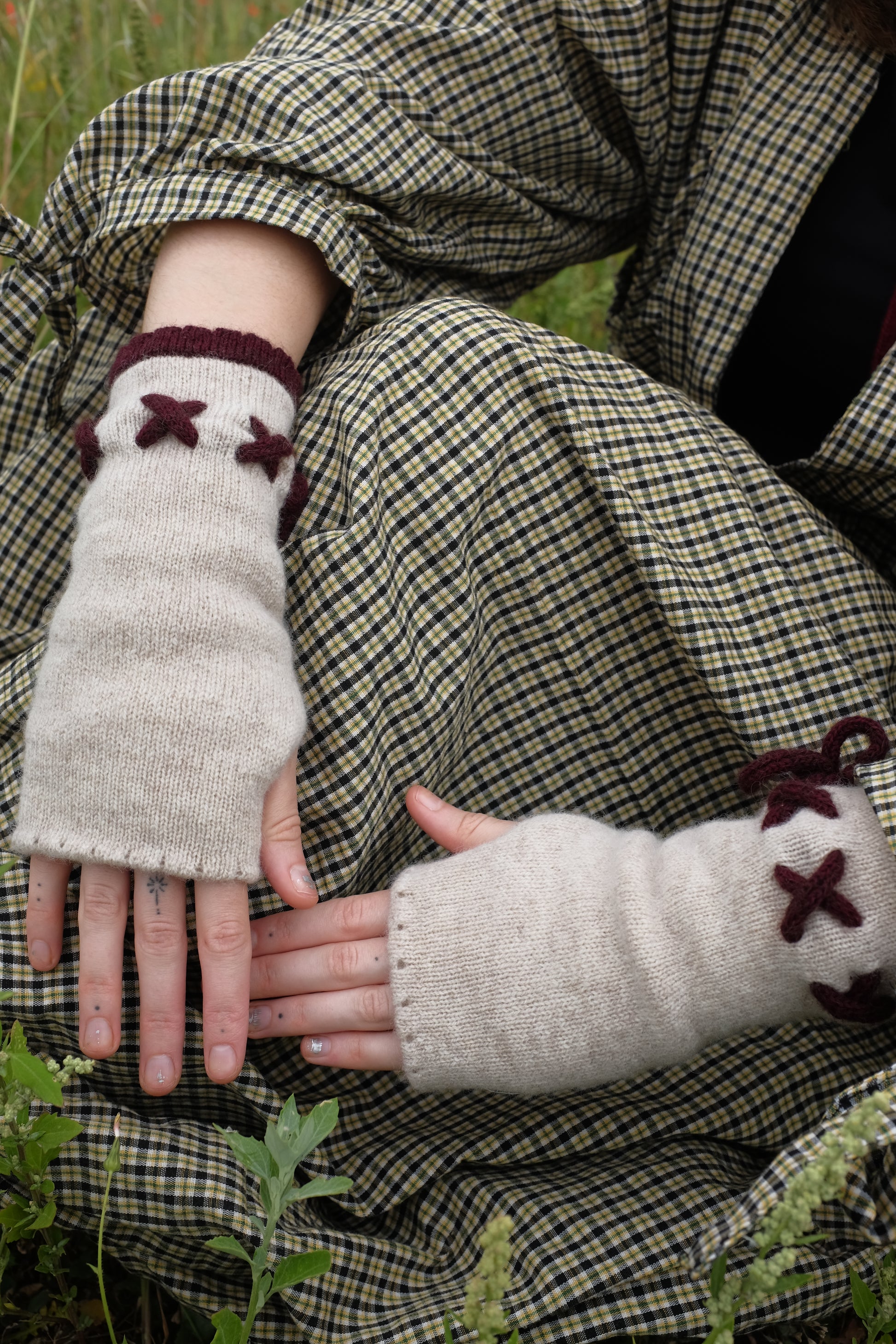 A woman sat in a filed, focuses only on her hands wearing a pair of fingerless gloves in plum and cream. The base of the gloves is cream with the tie detailing and trim in a plum. The background of the image is the woman’s trendy autumnal outfit. 