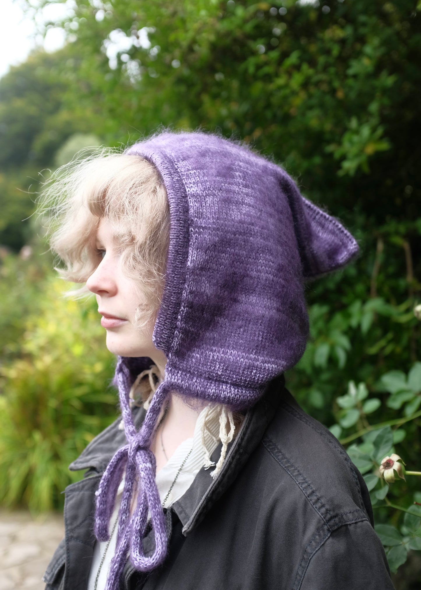 Person wearing a purple knitted hood outdoors with greenery in the background