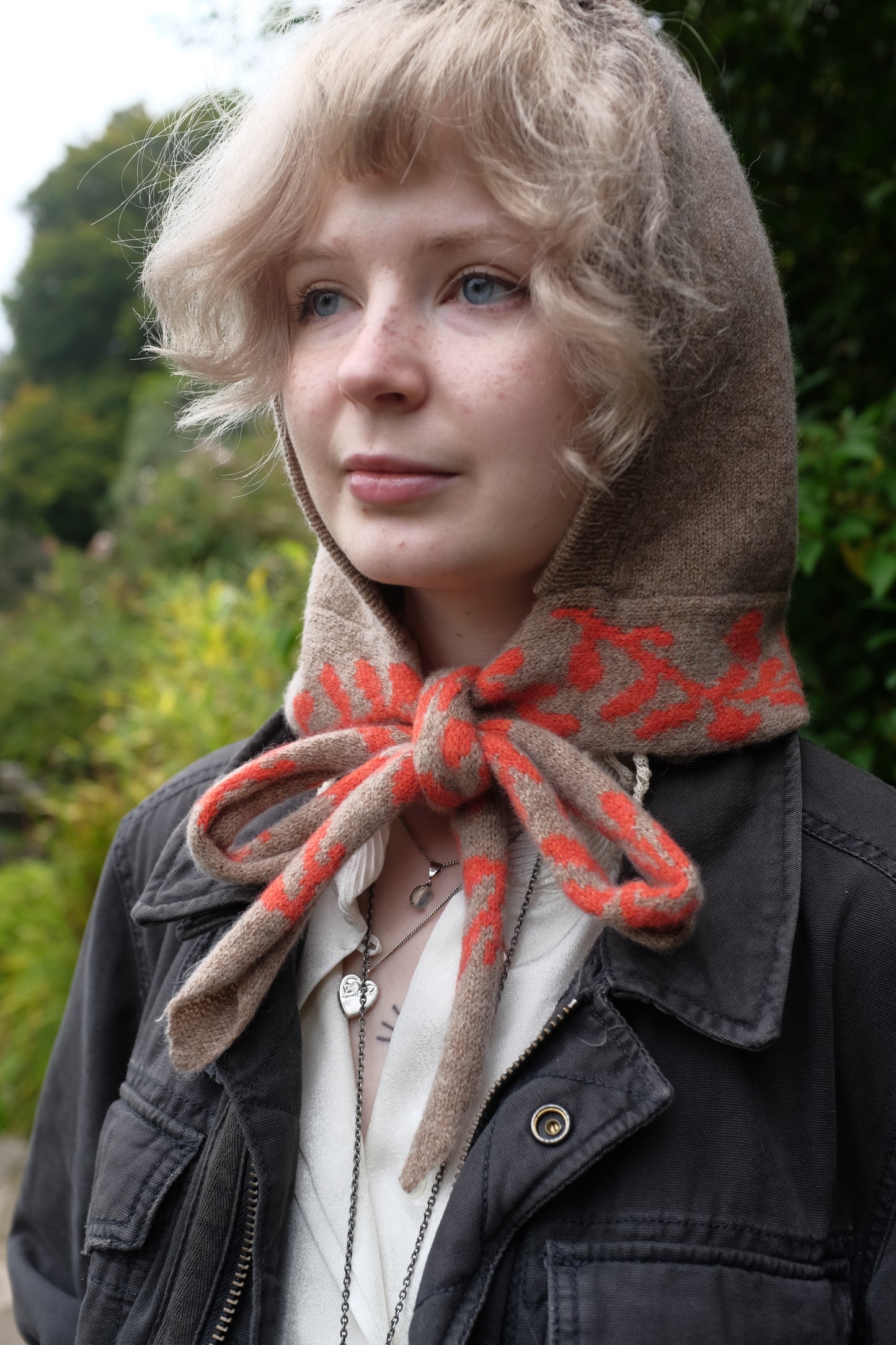 A front view of a woman wearing a lambswool knitted bonnet hood. The base of the bonnet is a tan colour. There is a fire orange pattern on the bottom of the bonnet tie that vis longer, mimicking a scarf. The pattern on the is a sequenced of laurel leaves and hearts. The women has tied the scarf in a bow and behind her is a mix of green autumnal leaves 