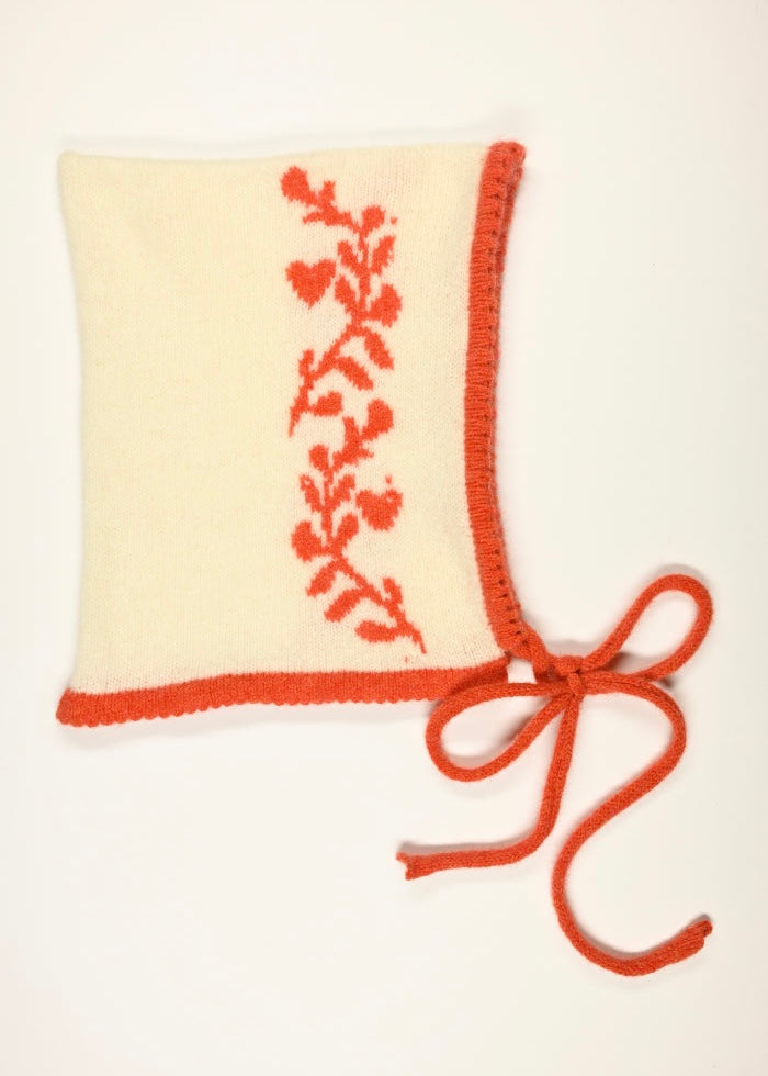 Knitted cream cashmere bonnet with an orange laurel leaf floral design and a bow on a white background