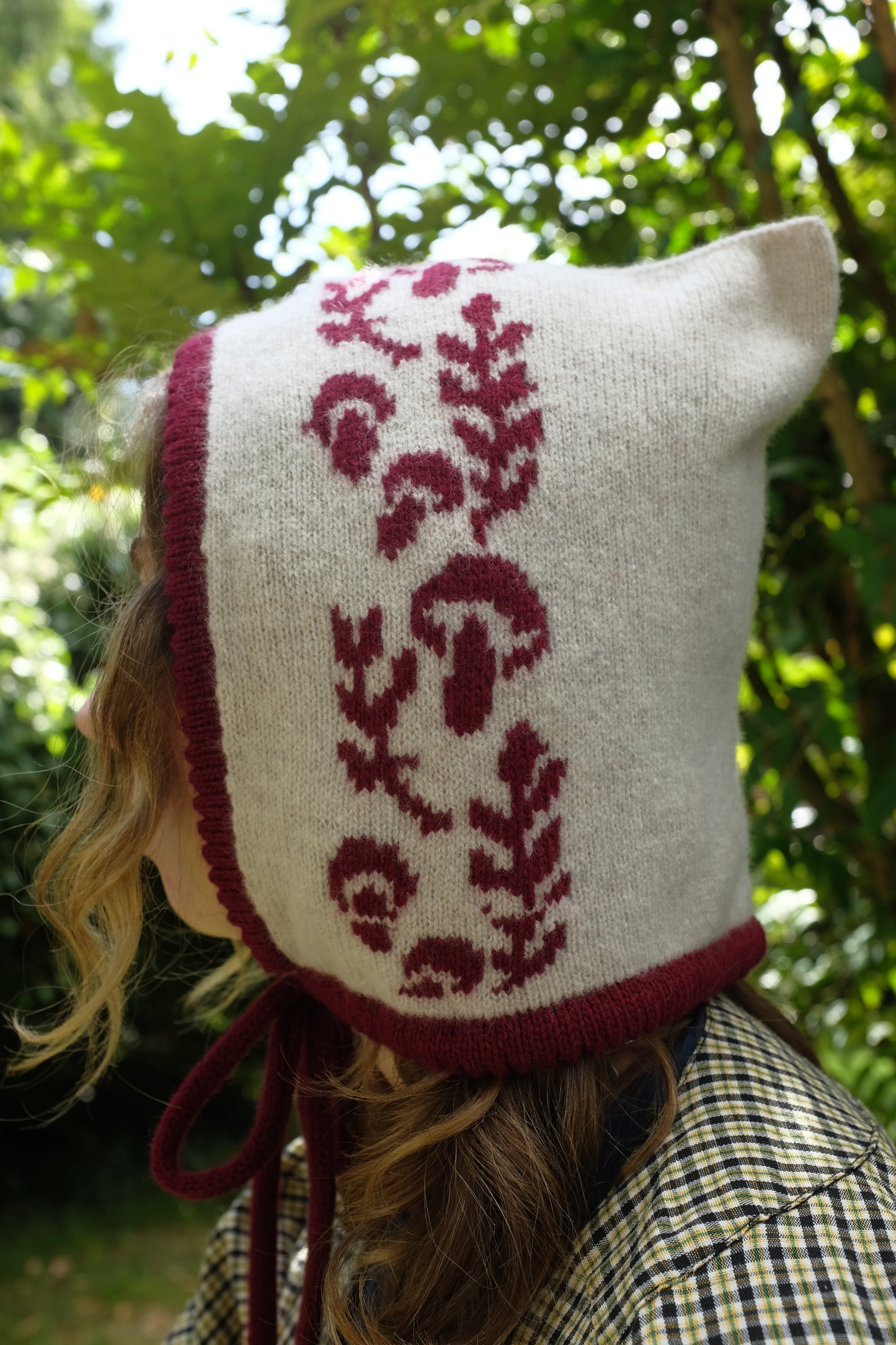 A close up of a women stood looking to the side wearing a cashmere and lambswool knitted bonnet. The base is cream with the trim, pattern and tie being a deep red. The pattern is made up of a range of mushrooms and leaves, scattered satisfyingly up the bonnet. Behind her are large green trees, highlighted by sunlight