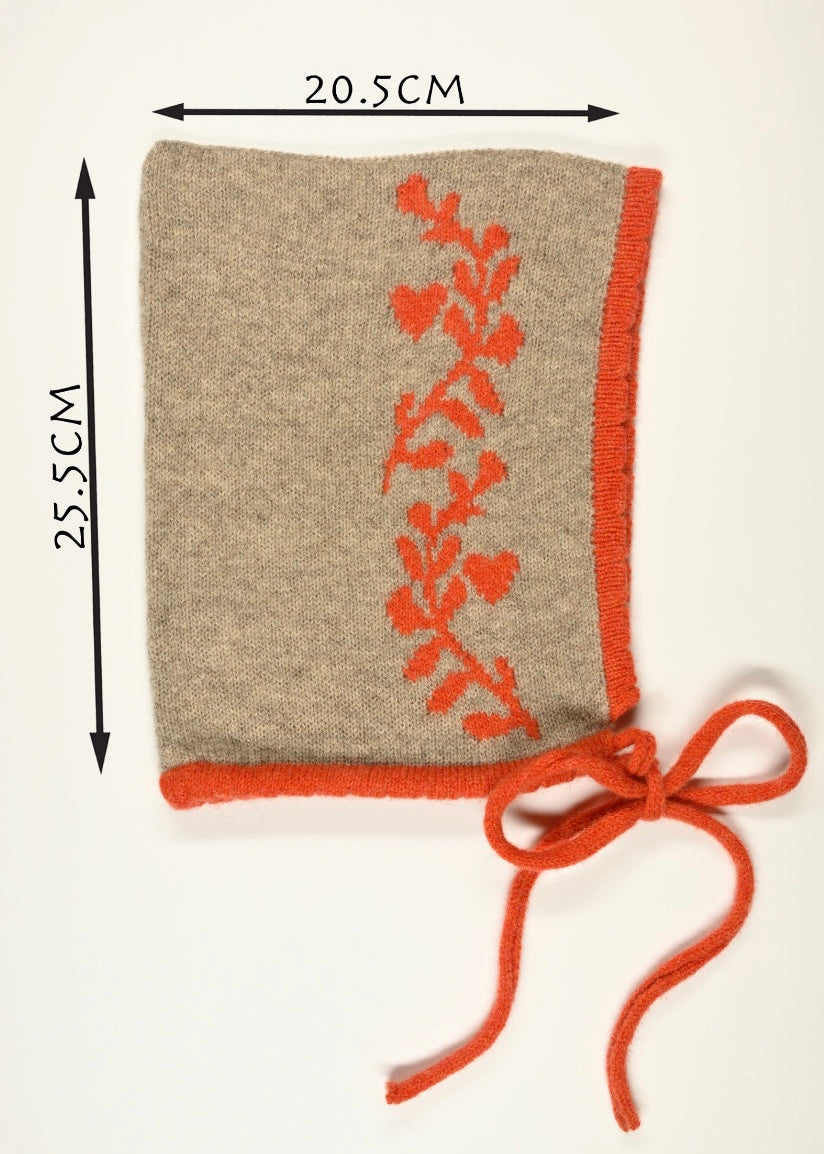 Tan knitted cashmere bonnet  with an orange laurel leaf design and a ribbon, shown with measurements (20.5cm wide by 25.5cm long) on a white background.