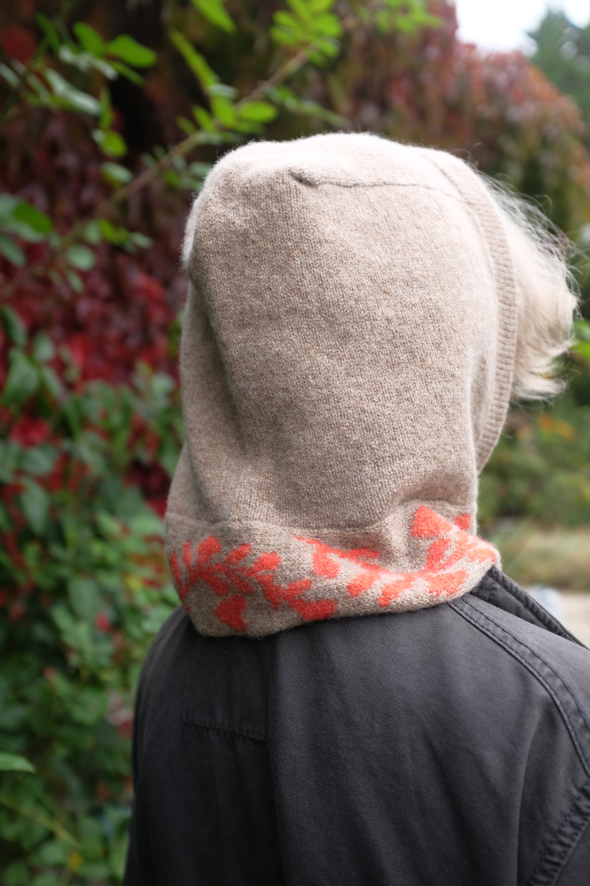 A back view of a woman wearing a lambswool knitted bonnet hood. The base of the bonnet is a tan colour. There is a fire orange pattern on the bottom of the bonnet tie that vis longer, mimicking a scarf. The pattern on the is a sequenced of laurel leaves and hearts. The women has tied the scarf loosely and behind her is a mix of green autumnal leaves 