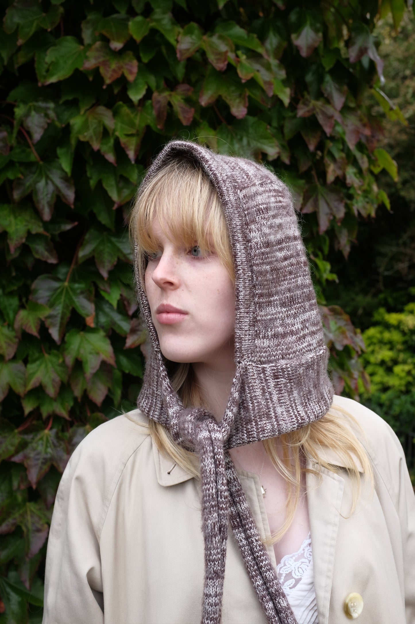 Person wearing a burgundy mohair bonnet-style hood that fully covers the head and neck. With two long, soft extensions hanging down from the front. The hood is thick and textured, offering warmth and a cosy appearance, ideal for cold weather. The person is dressed in a light coloured coat and stands outdoors against a backdrop of green and red foliage 