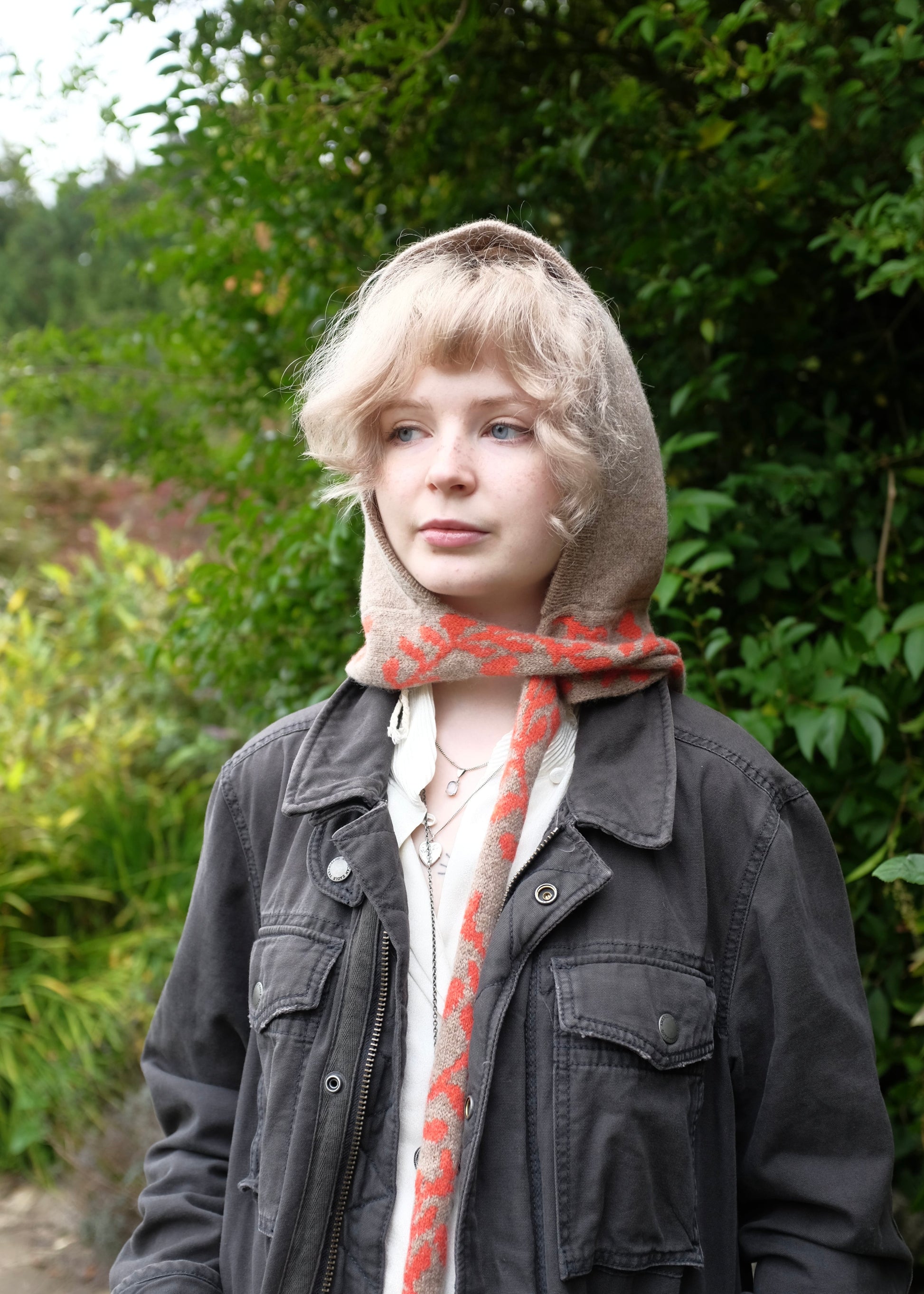 Person wearing a knitted lambswool hooded bonnet in tan with a orange scarf detailing in a natural setting