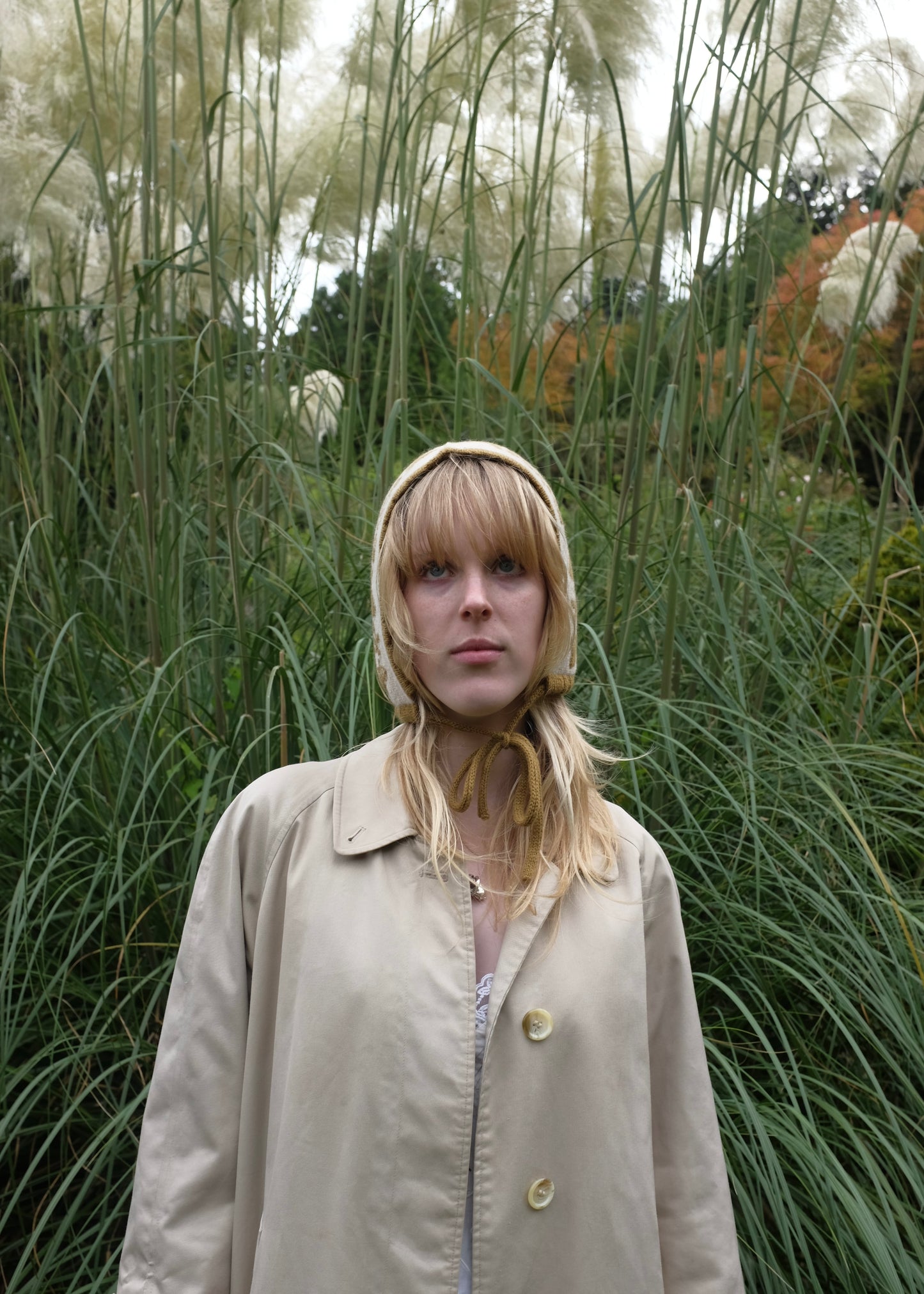 Person wearing a beige coat and hood in a natural setting with tall grasses.