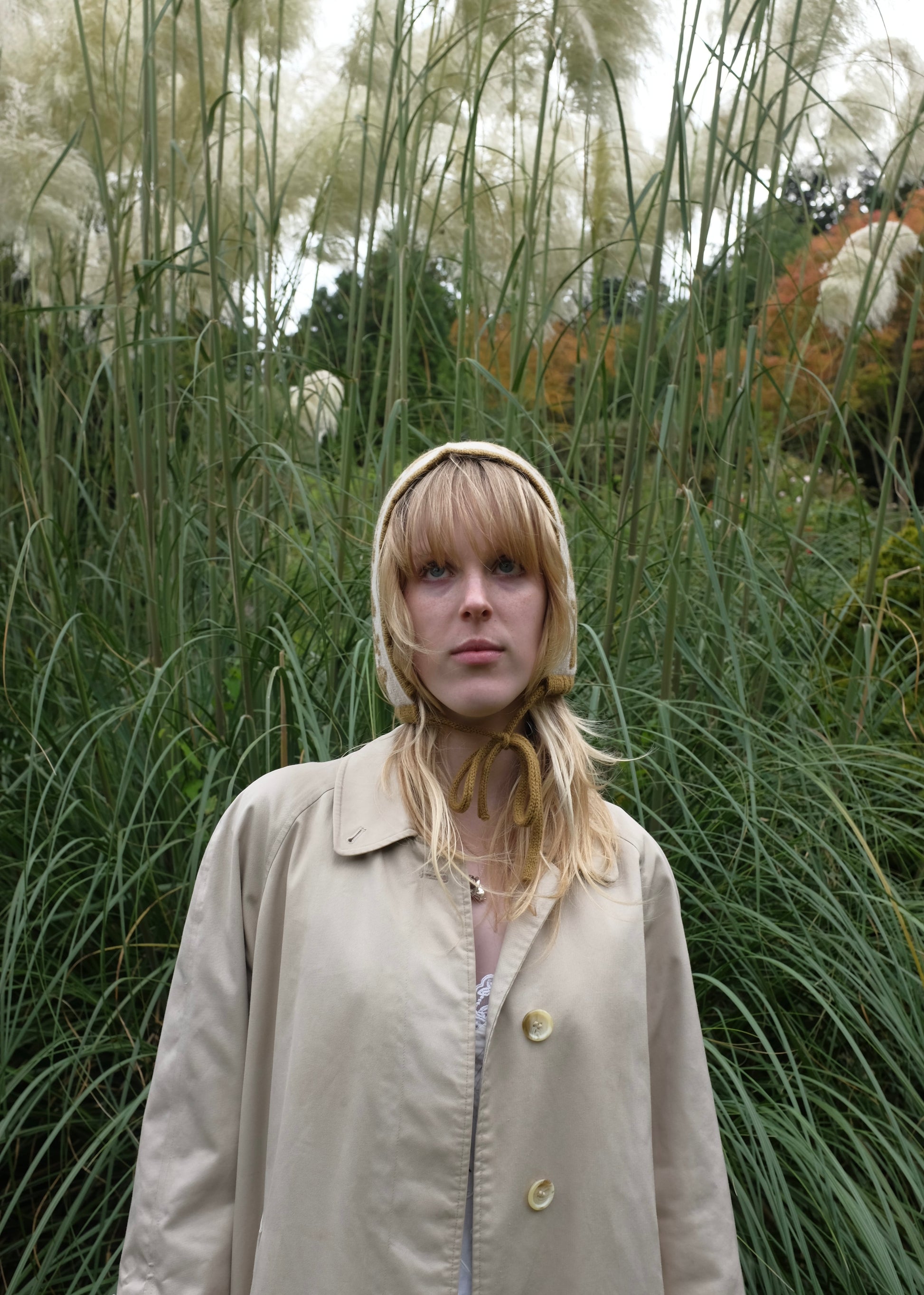 Person wearing a beige coat and hood in a natural setting with tall grasses.