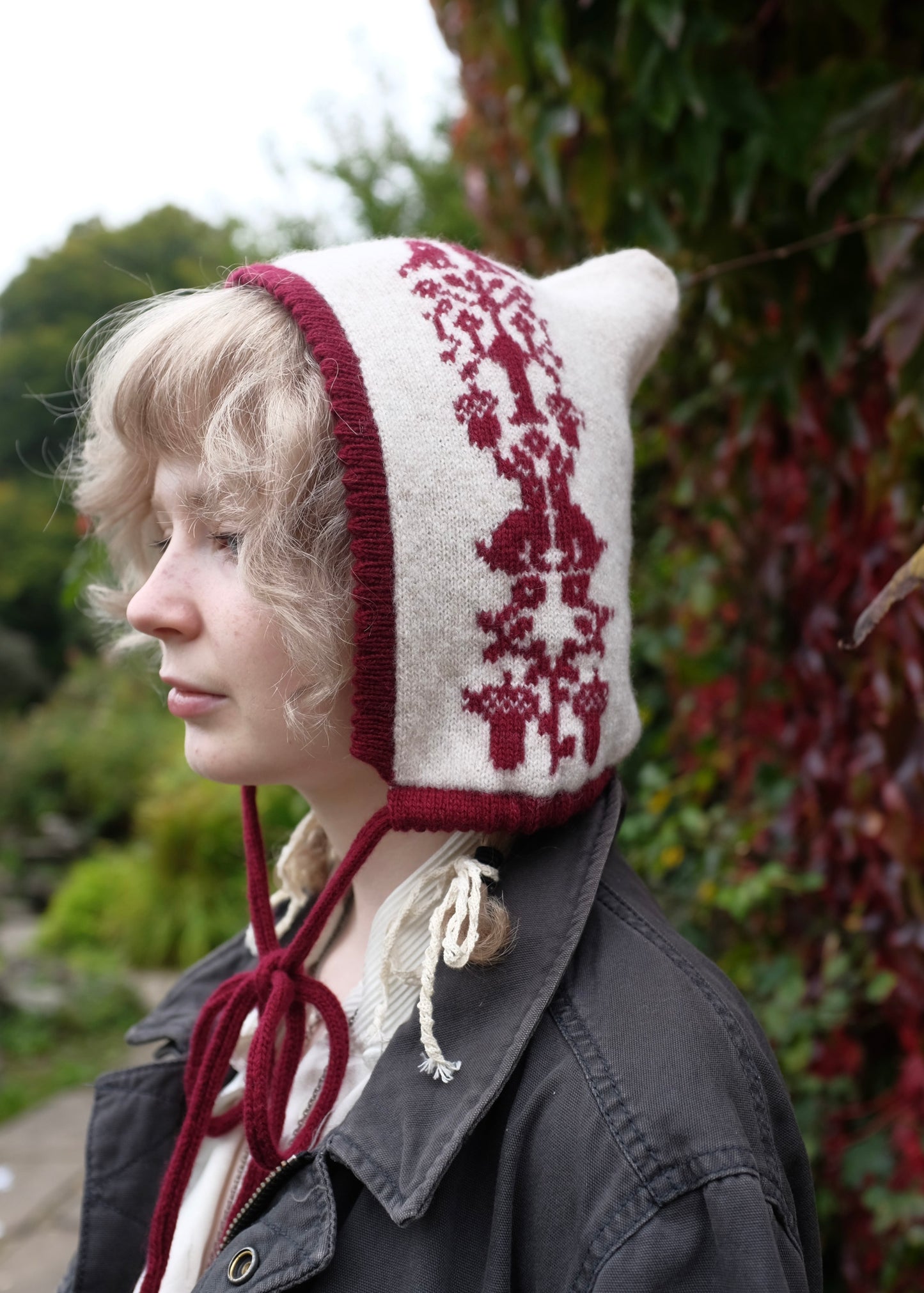 Person wearing a cream knitted bonnet with red patterns and trim outdoors.
