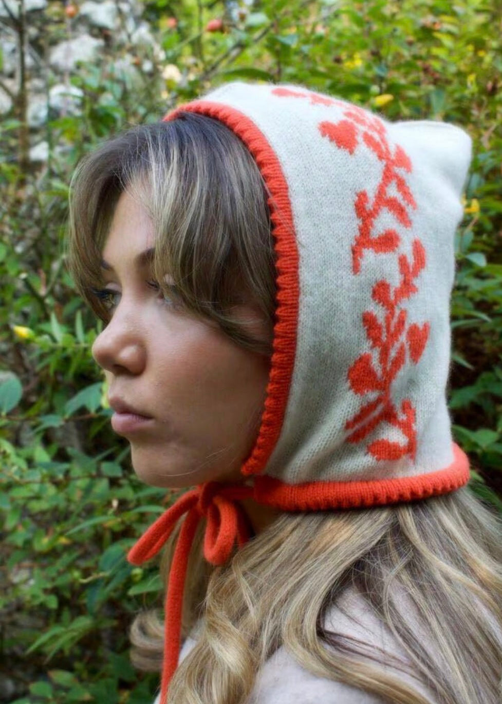 Person wearing a white hood with red patterns and orange trim in a natural setting