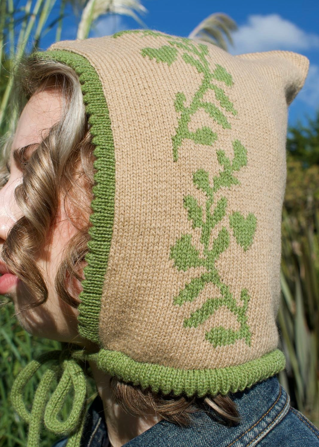 Person wearing a tan knitted hood with green trim and laurel leaf pattern, standing outdoors.