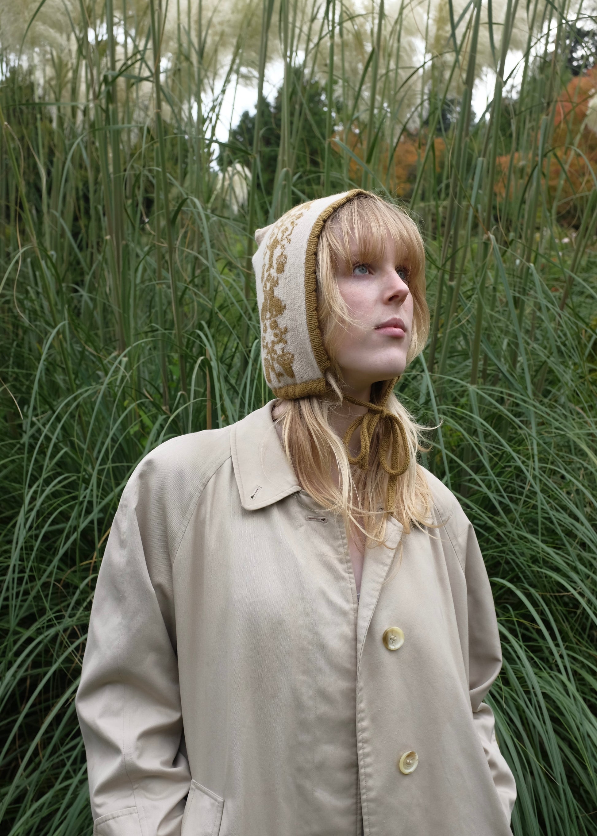 Person wearing a beige coat and bonnet in a grassy outdoor setting