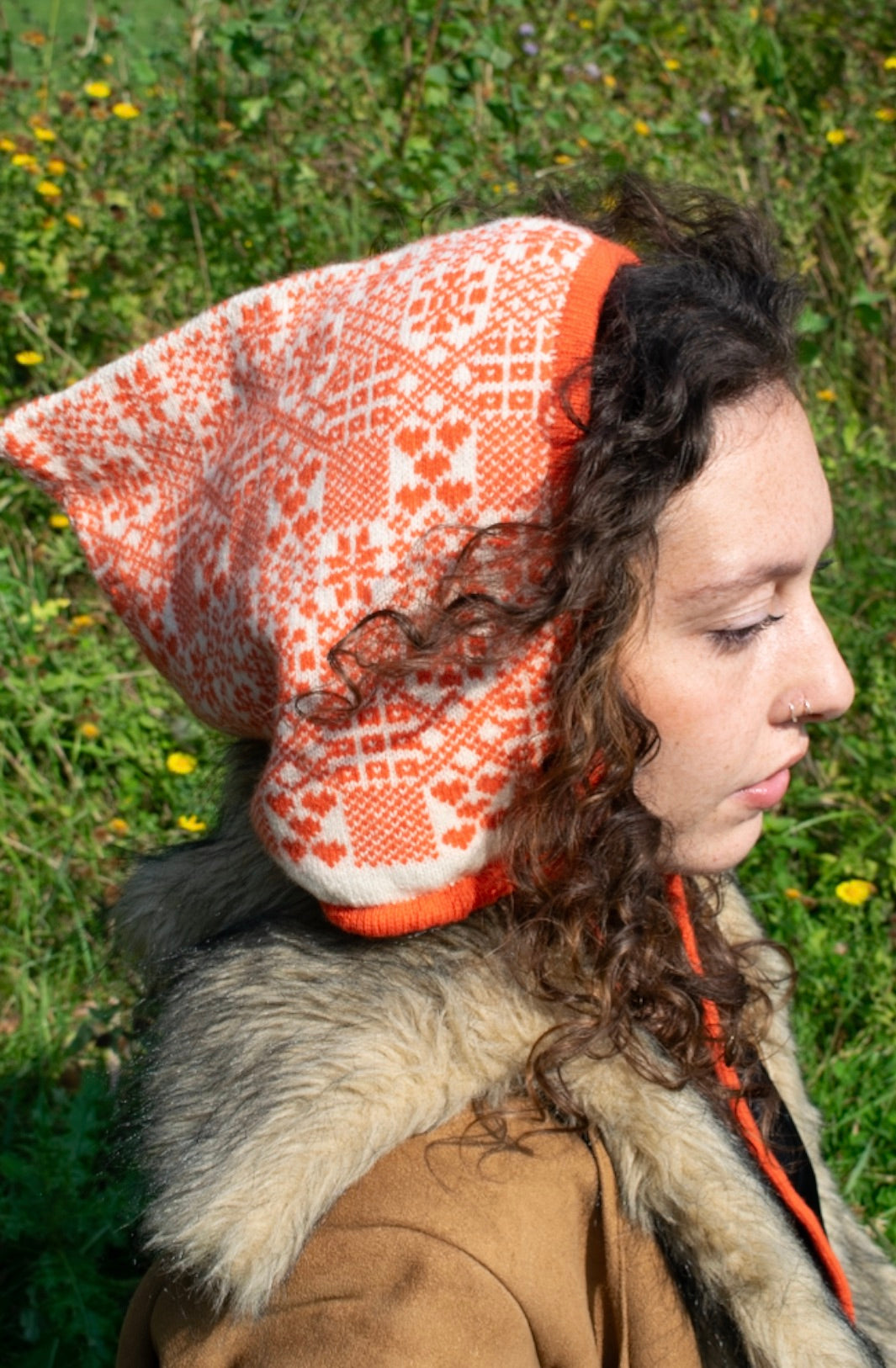 A person is outdoors side view , surrounded by tall grass and tress, wearing a hand-knitted cashmere hood bonnet. The hood features a symmetrical orange and white pattern inspired by traditional Nordic or Fair isle designs. It ties neatly under thr chin with an orange string, showcasing detailed craftsmanship and cultural textile artistry. 