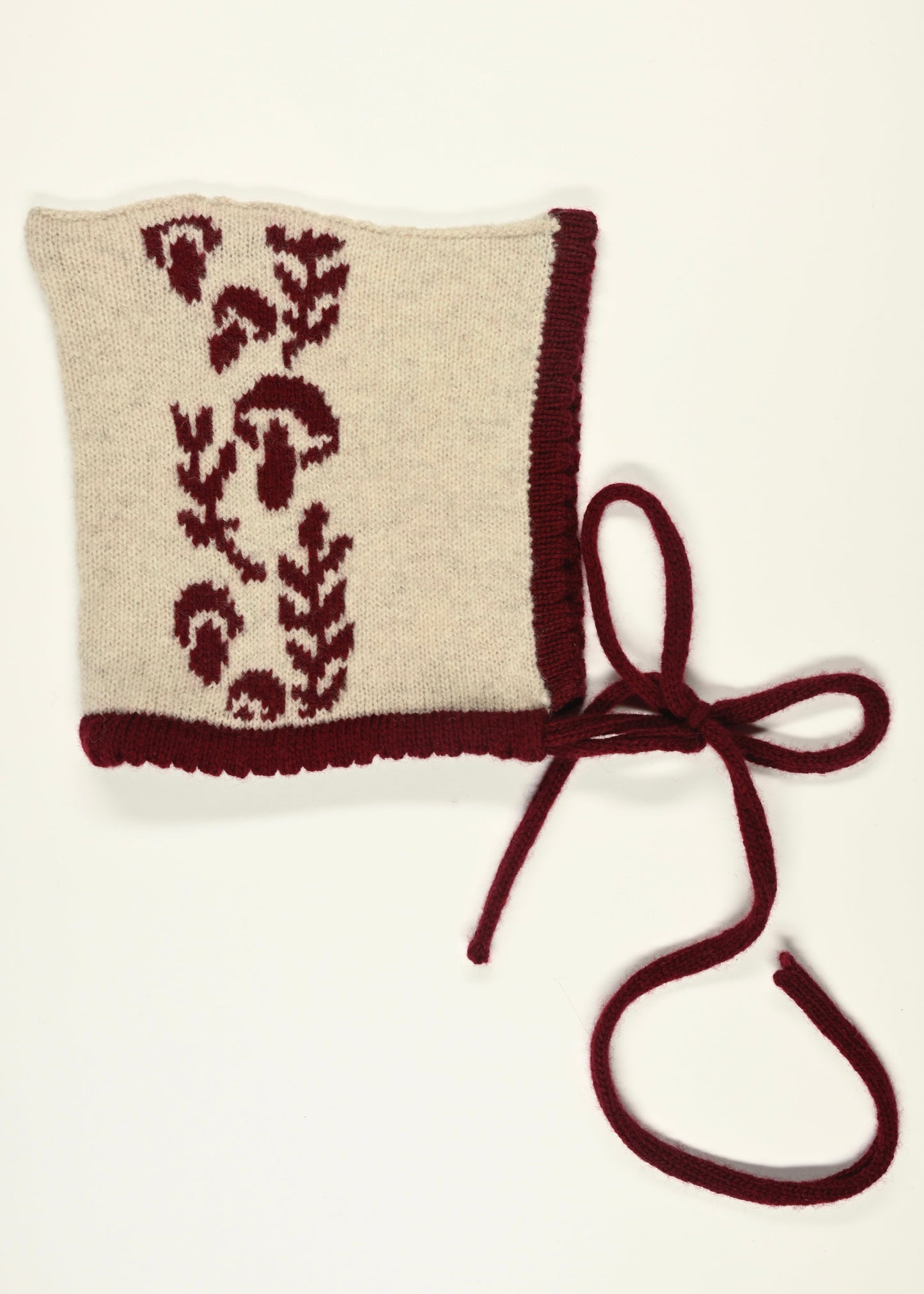 Knitted baby bonnet with floral mushroom pattern and burgundy strap on a white background