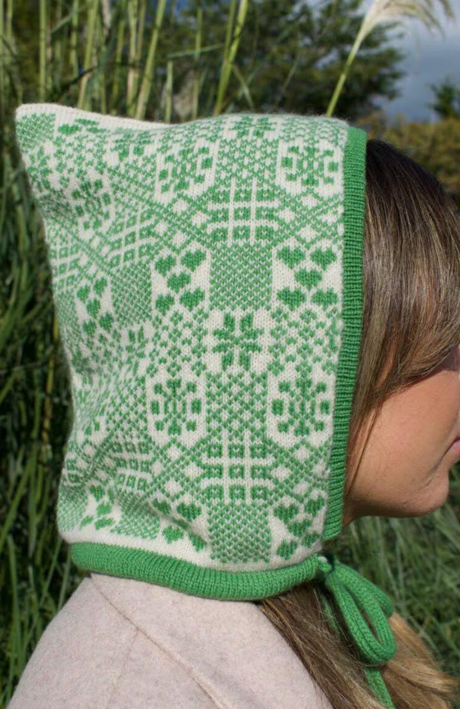 A person is outdoors pictured from a side view, surrounded by tall grass and tress, wearing a hand-knitted cashmere hood bonnet. The hood features a symmetrical green and white pattern inspired by traditional Nordic or Fair isle designs. It ties neatly under thr chin with a green string, showcasing detailed craftsmanship and cultural textile artistry. 