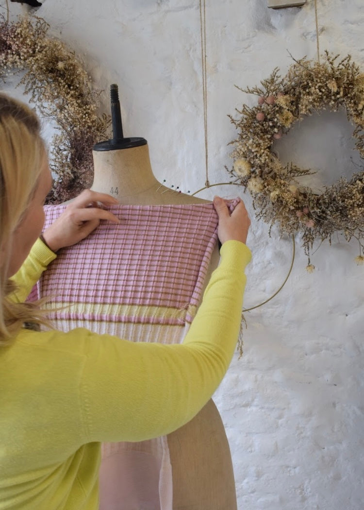 Person adjusting a dress form with a checkered garment against a white wall with wreaths.