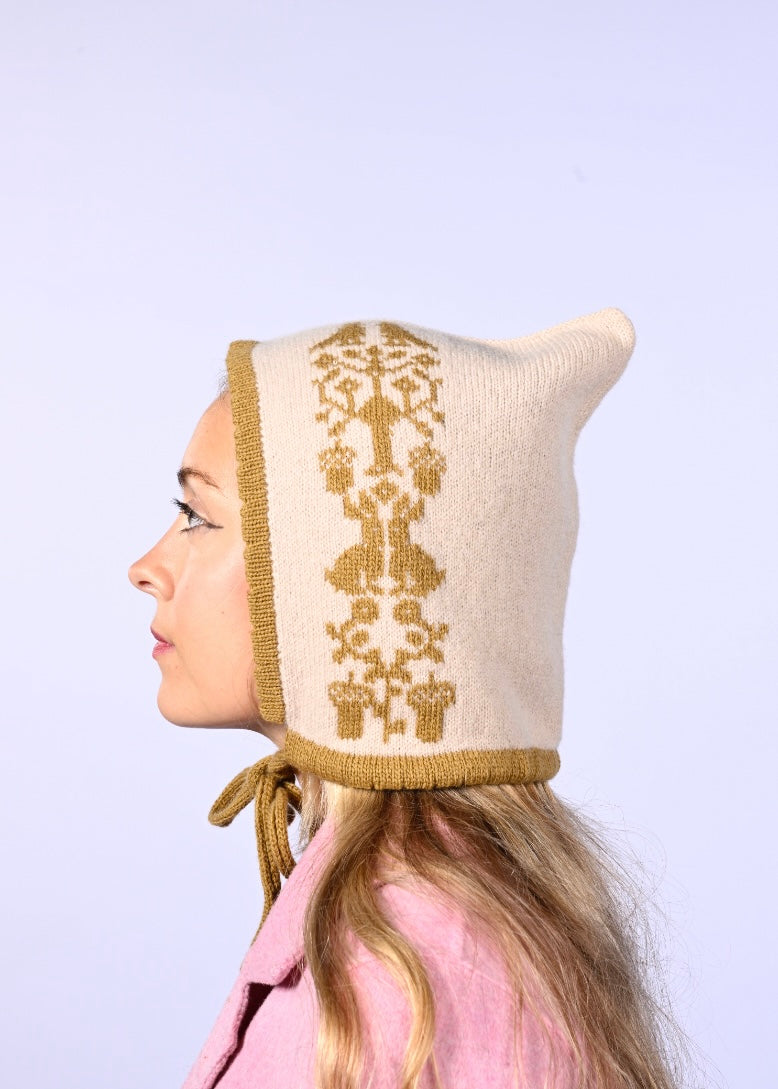 Person wearing a cream-colored knitted bonnet with gold embroidery on a light purple background