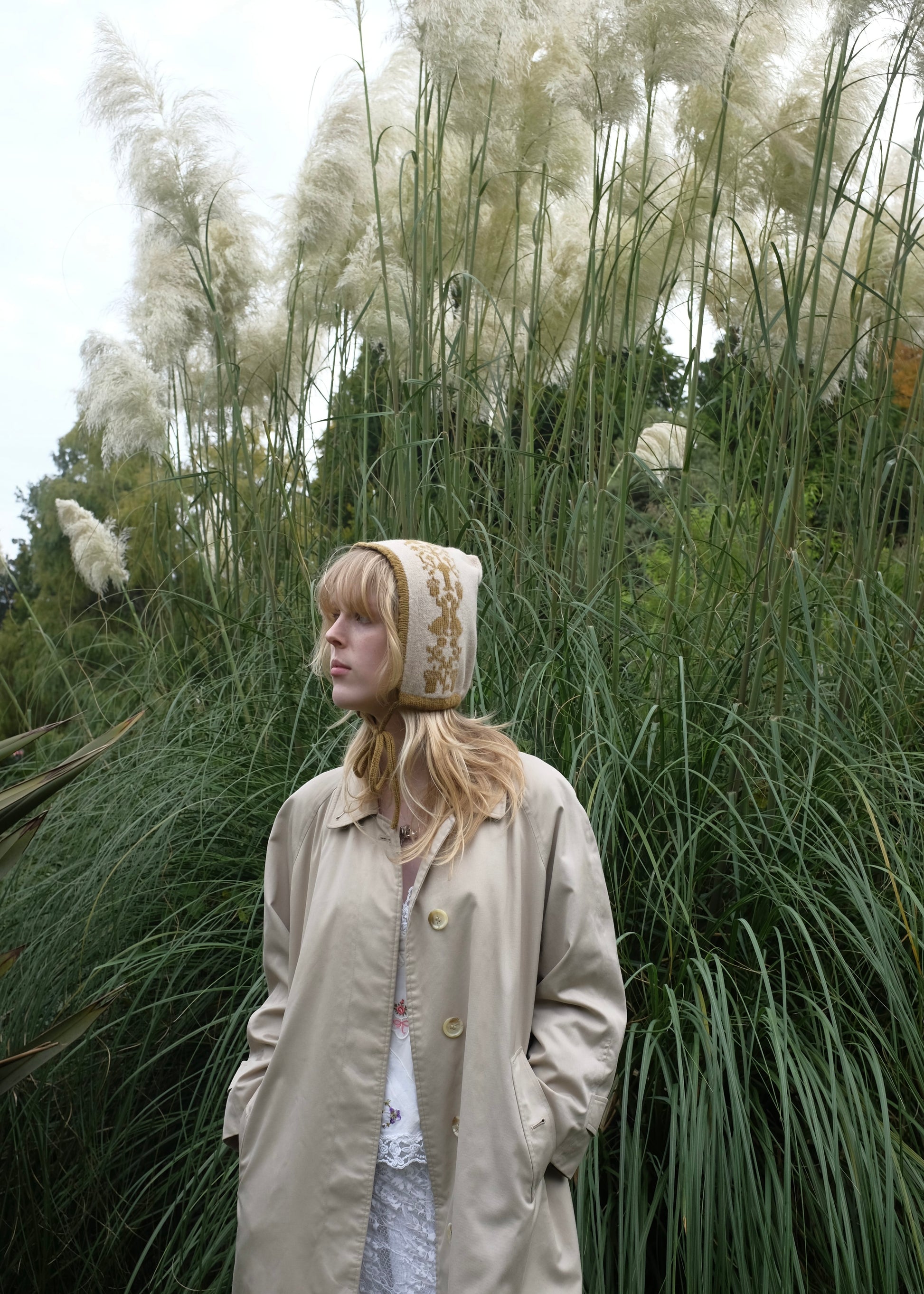 Woman wearing a beige coat and bonnet standing among tall grasses