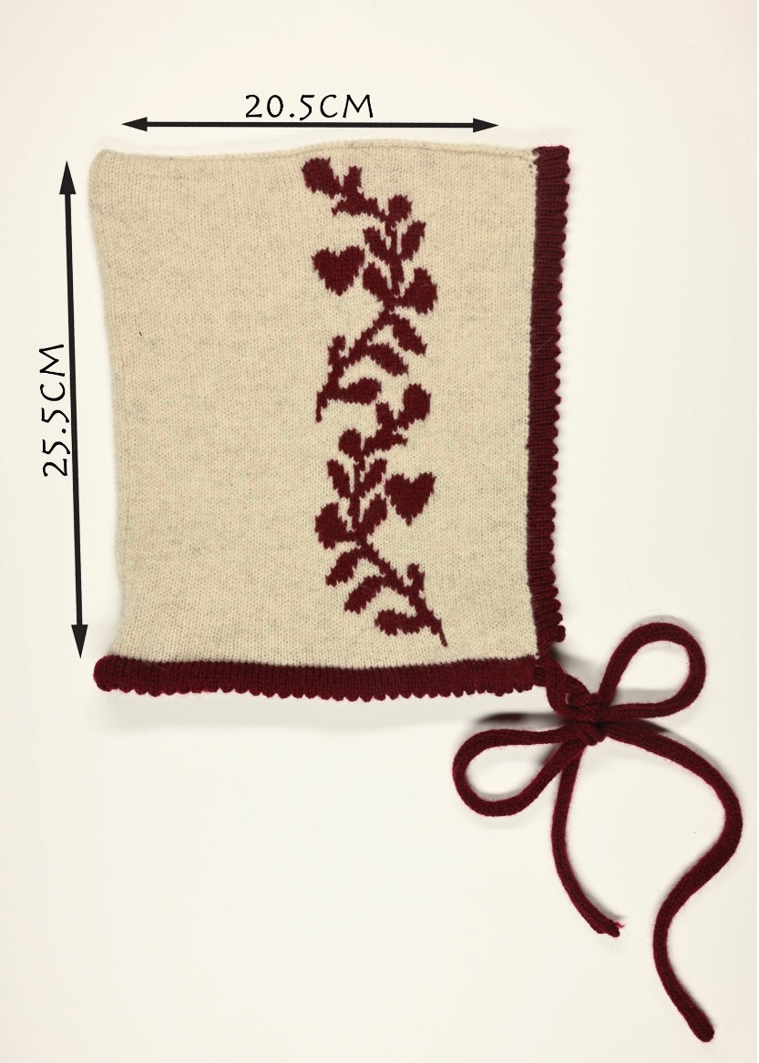 A cream bonnet with red laurel leaf floral design and a bow, shown with measurements (20.5cm wide by 25.5cm long) on a beige background