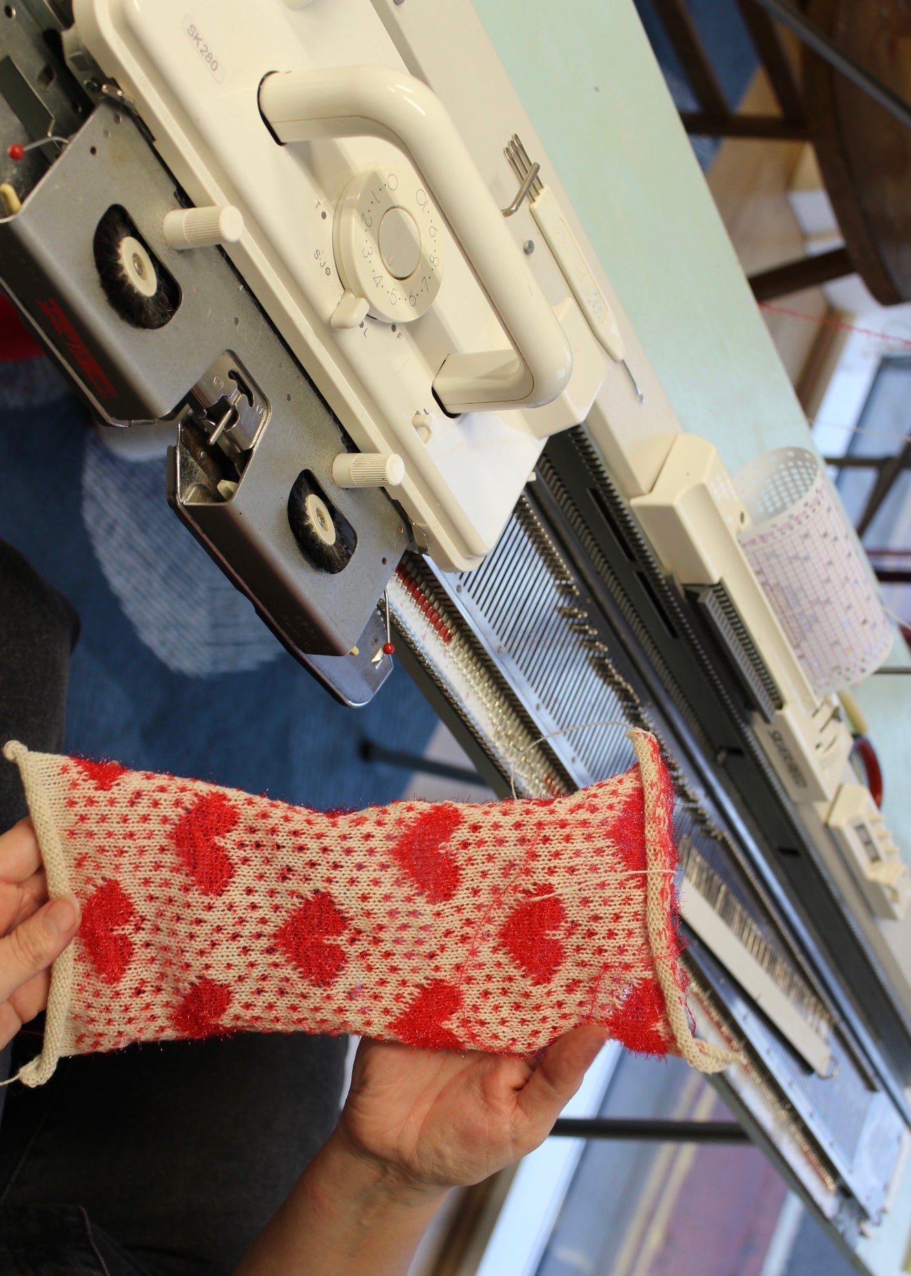 Person holding a red and white patterned in front of a knitting machine.