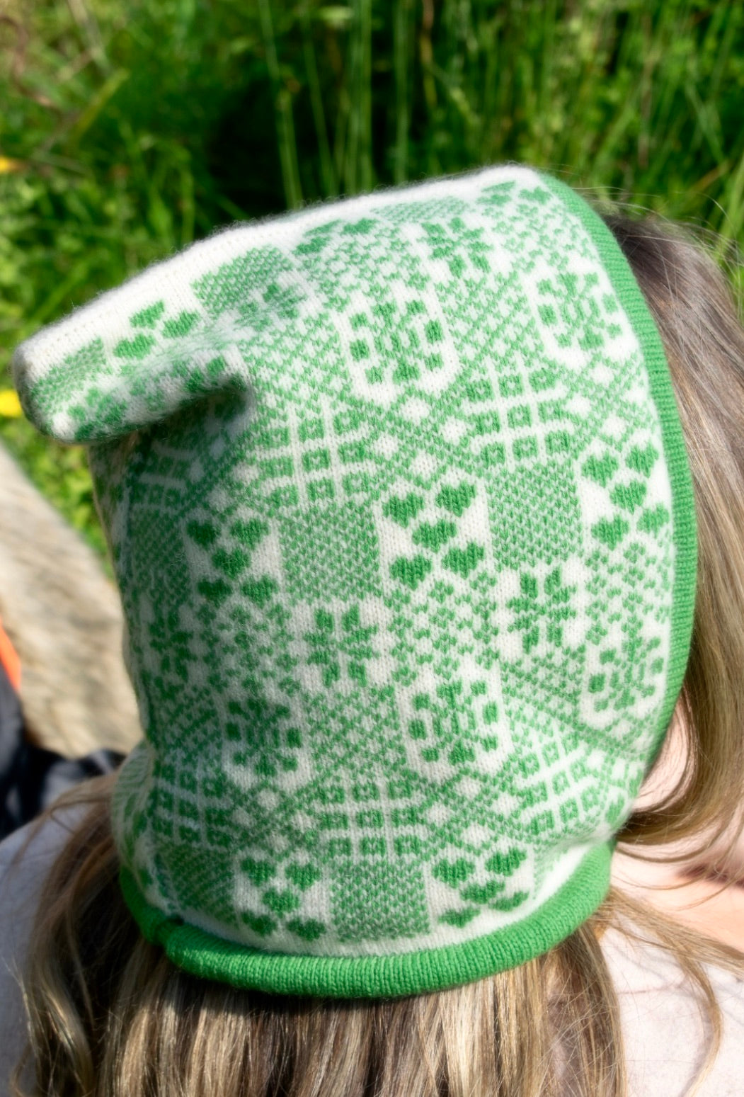 A person is outdoors pictured from behind , surrounded by tall grass and tress, wearing a hand-knitted cashmere hood bonnet. The hood features a symmetrical green and white pattern inspired by traditional Nordic or Fair isle designs. It ties neatly under thr chin with a green string, showcasing detailed craftsmanship and cultural textile artistry. 