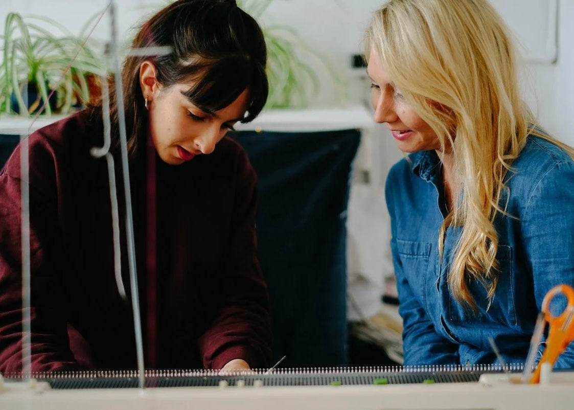 Two women sitting together a student and teacher , at a knit machine, with a neutral background.