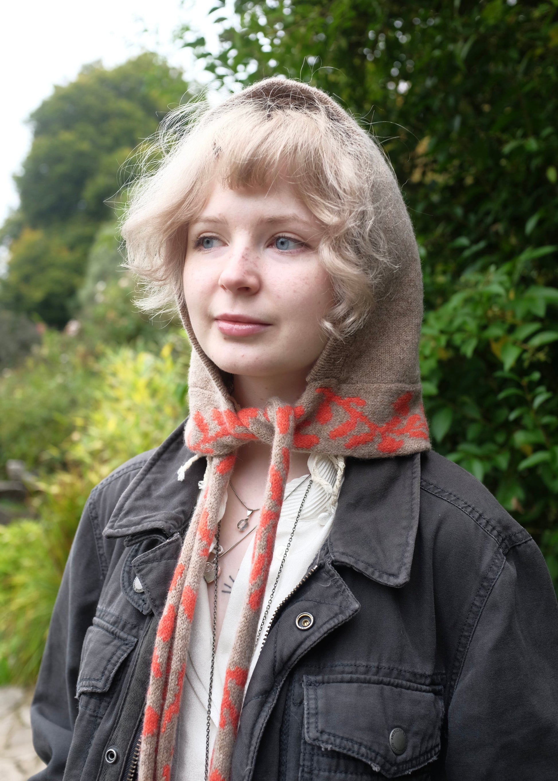 Person wearing a tan hood Bonet with a patterned scarf detailing of a orange laurel leaf in an outdoor setting