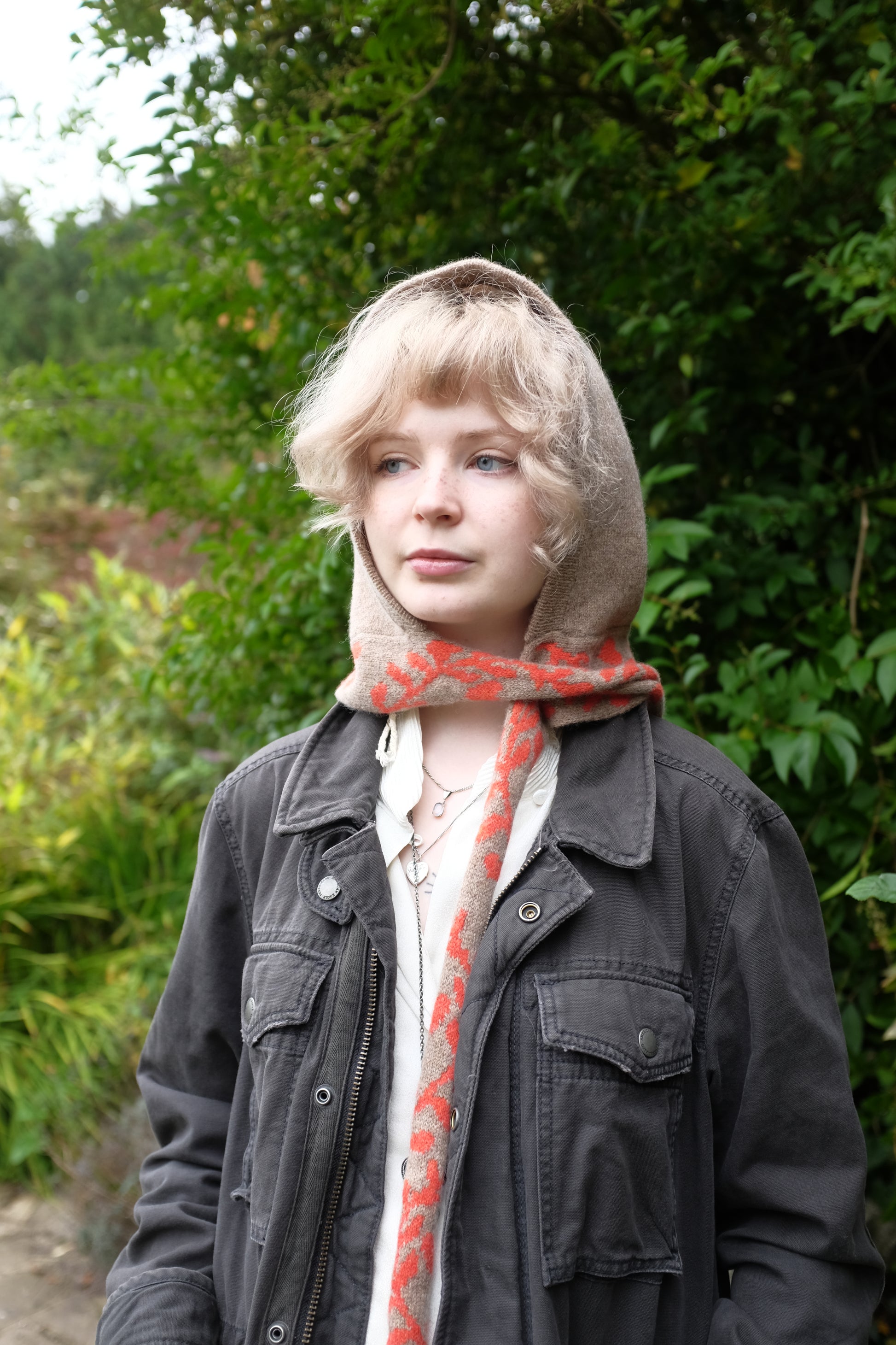 A front view of a woman wearing a lambswool knitted bonnet hood. The base of the bonnet is a tan colour. There is a fire orange pattern on the bottom of the bonnet tie that vis longer, mimicking a scarf. The pattern on the is a sequenced of laurel leaves and hearts. The women has draped the scarf around her neck and over her shoulder and behind her is a mix of green autumnal leaves 