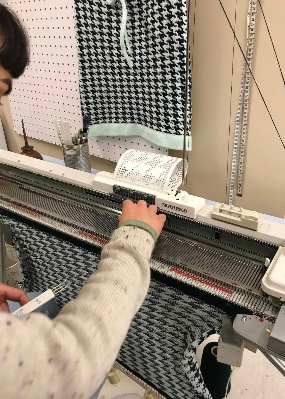 Person operating a knitting machine with a pattern guide in a workshop setting.