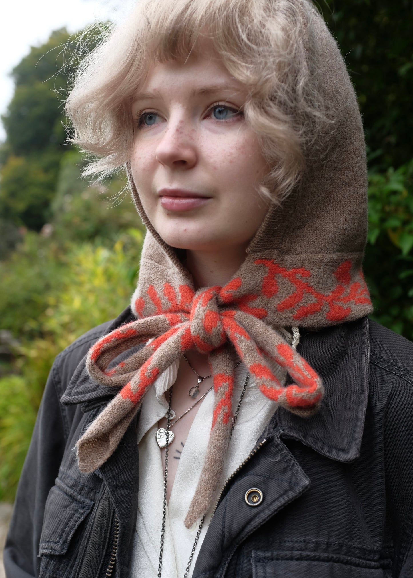 Woman wearing a knitted hooded bonnet in tan  with a red and beige patterned scarf detailing outdoors 