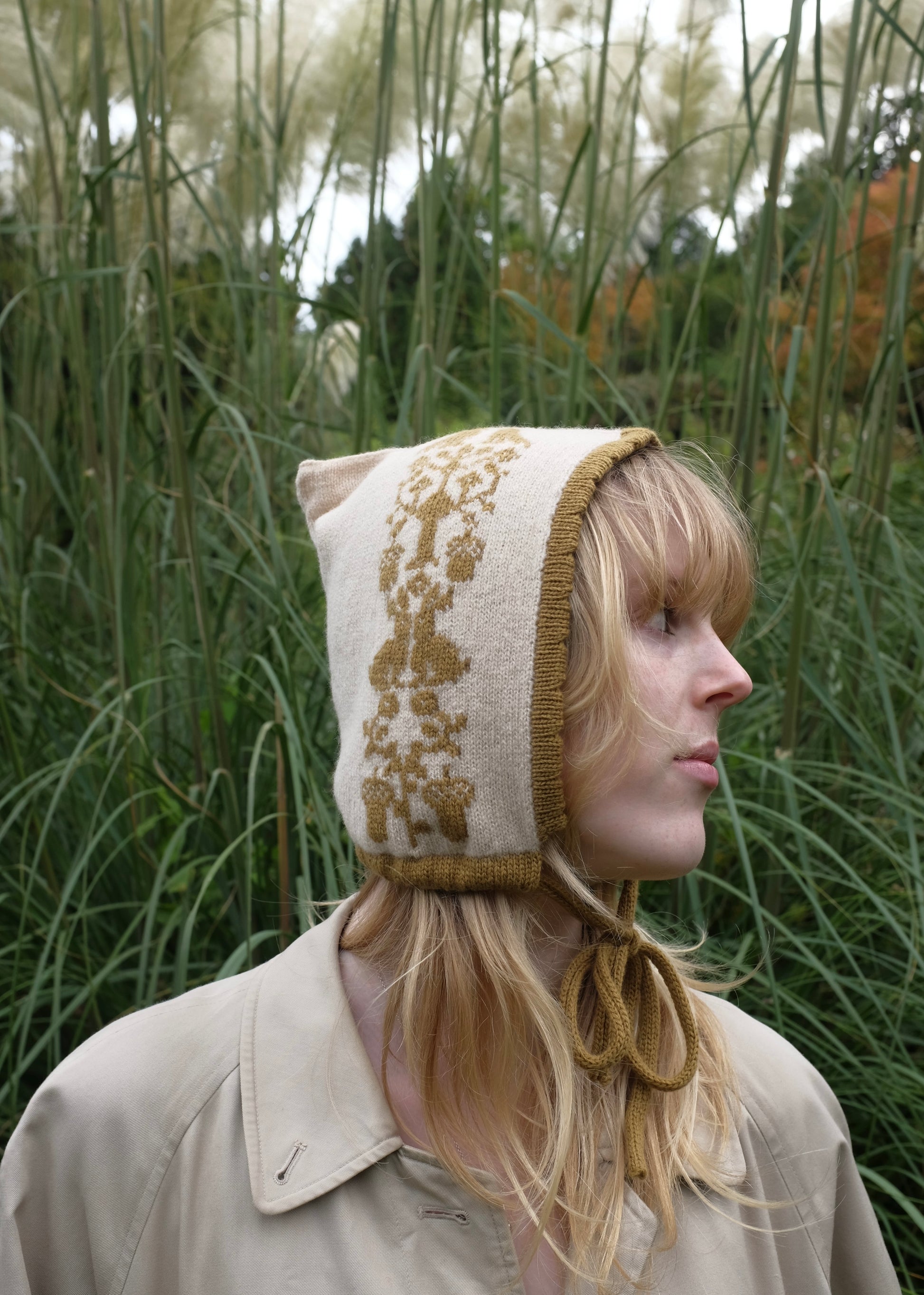 Person wearing a beige knitted bonnet with golden animal pattern, trim and tie in a natural setting
